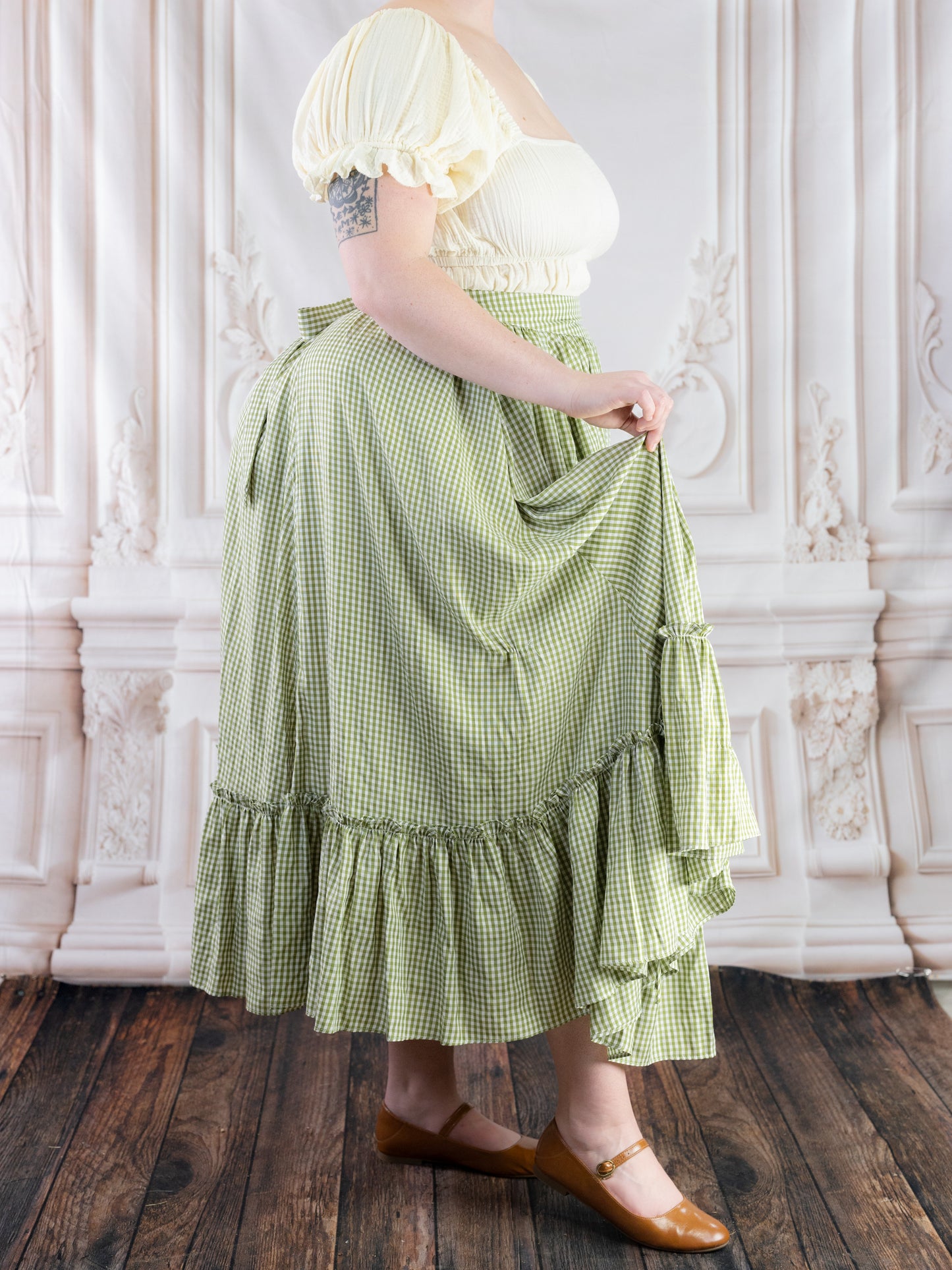 Woman wearing a green gingham ruffle skirt paired with a cream puff sleeve top and brown shoes in a vintage room setting