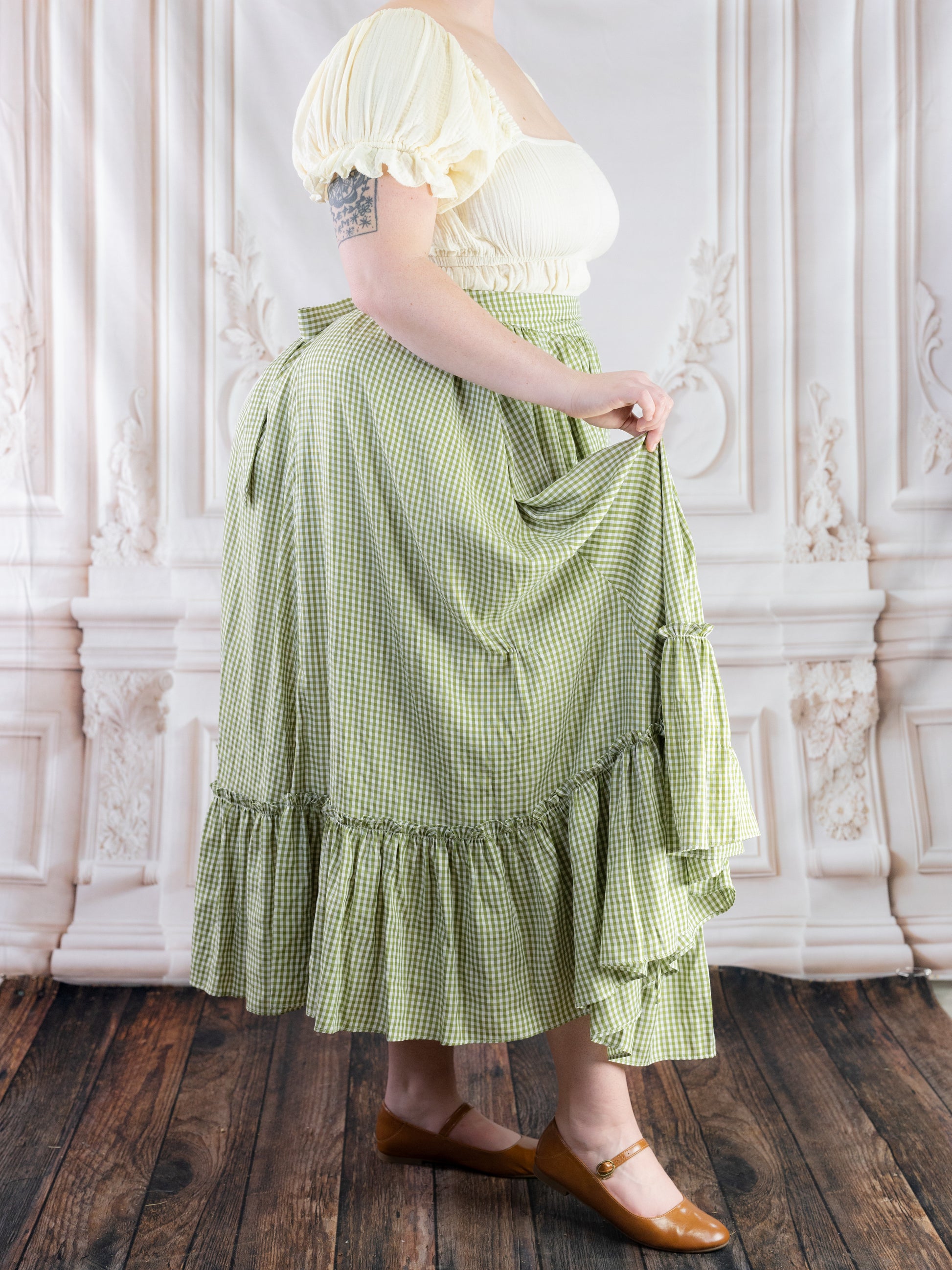 Woman wearing a green gingham ruffle skirt paired with a cream puff sleeve top and brown shoes in a vintage room setting