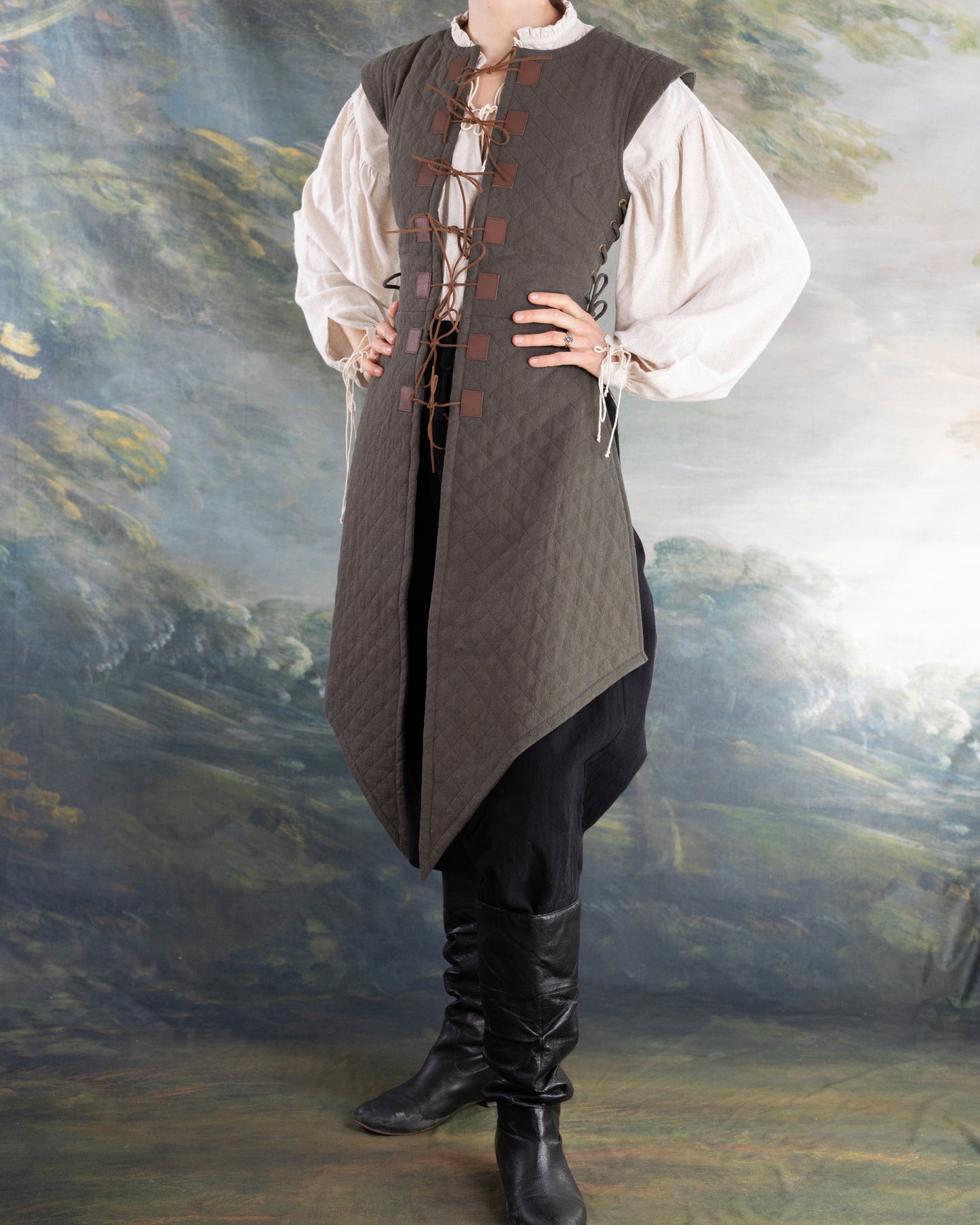 Woman wearing a dark gray quilted gambeson over a white shirt with black pants and boots standing in front of a painted backdrop