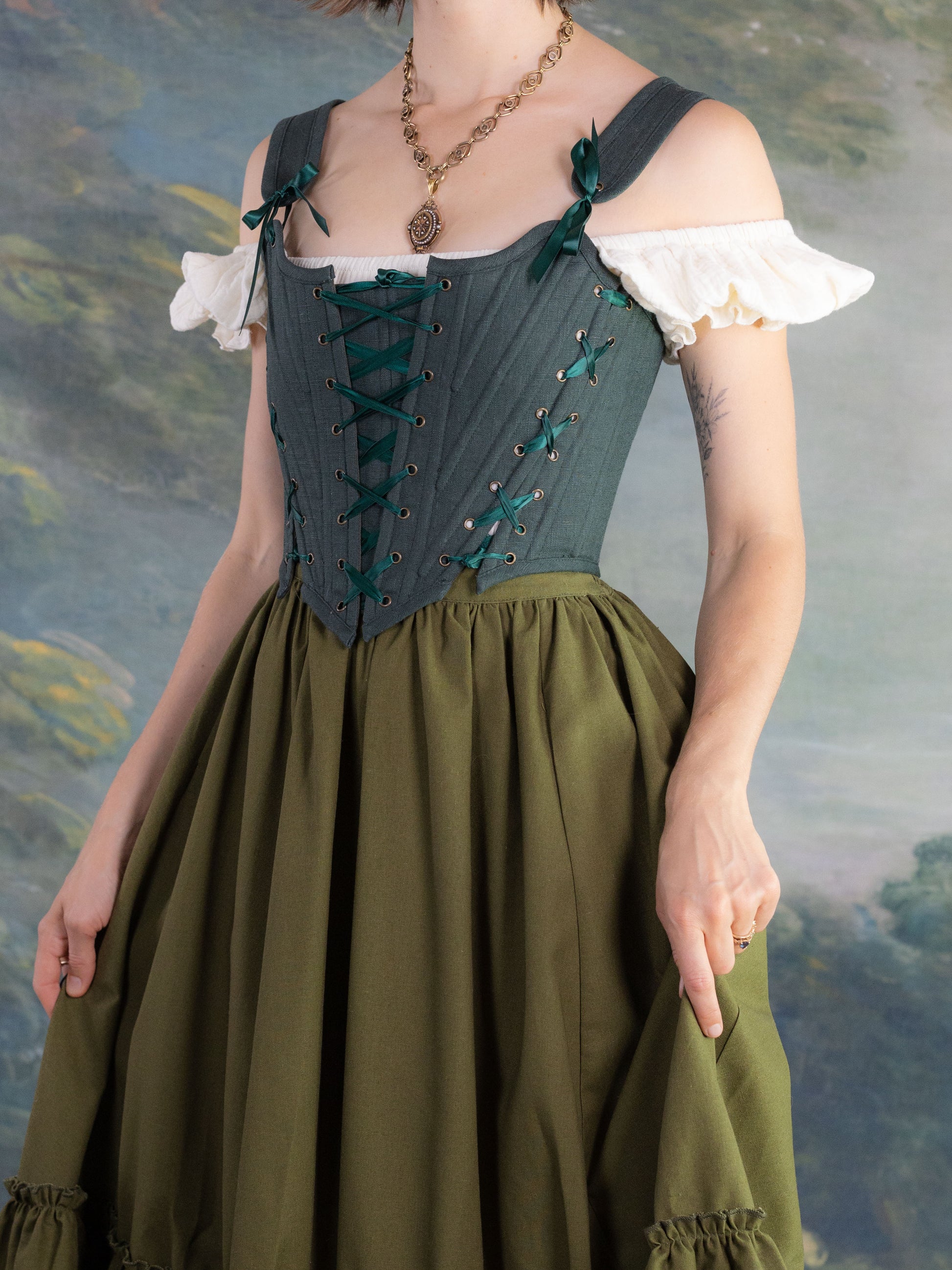 Woman wearing Forest Green Princess Stays corset with off-the-shoulder white blouse and olive green skirt in soft natural light