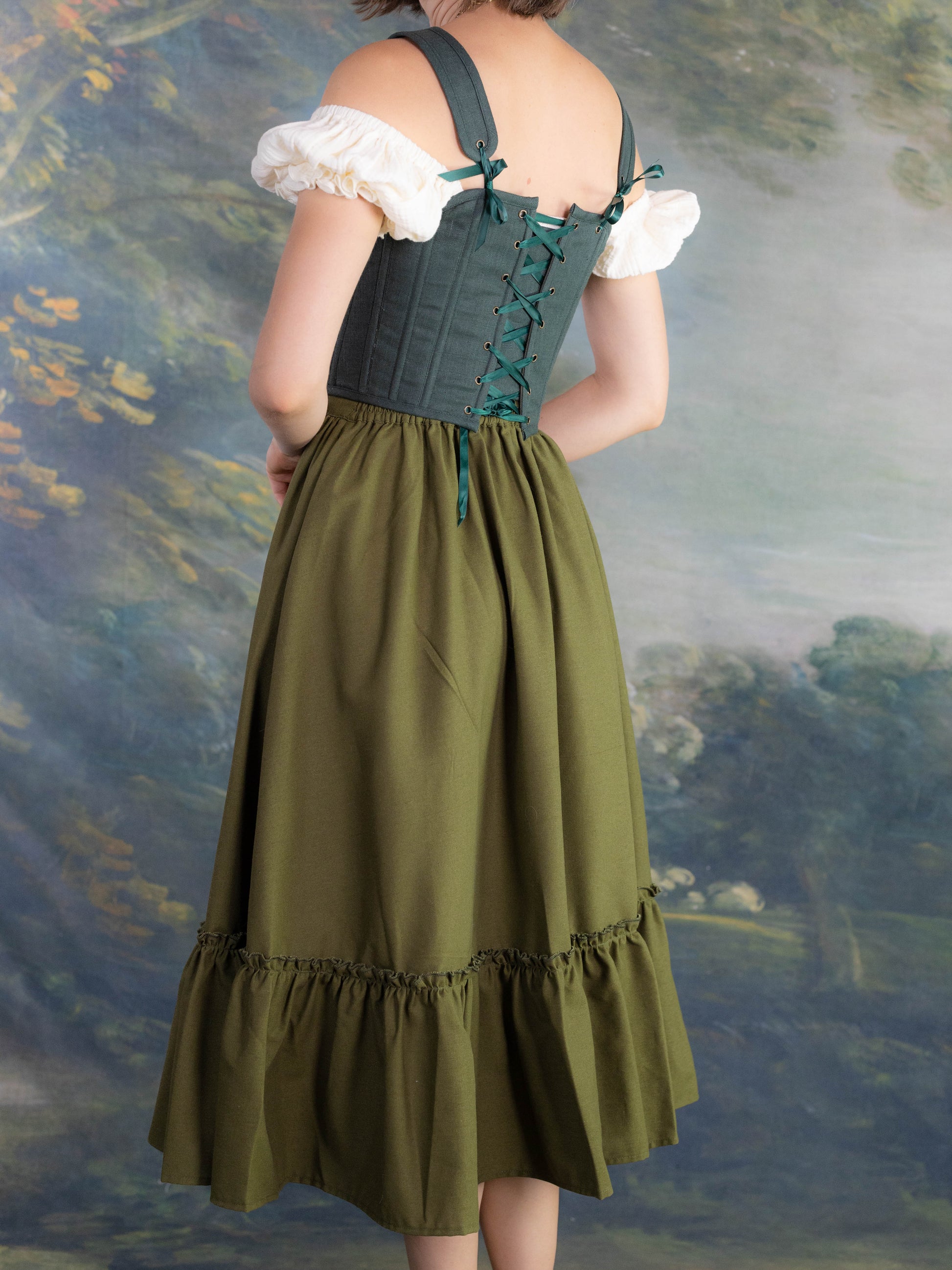 Back view of a woman wearing a Forest Green Princess Stays corset with a green skirt and white blouse in soft lighting