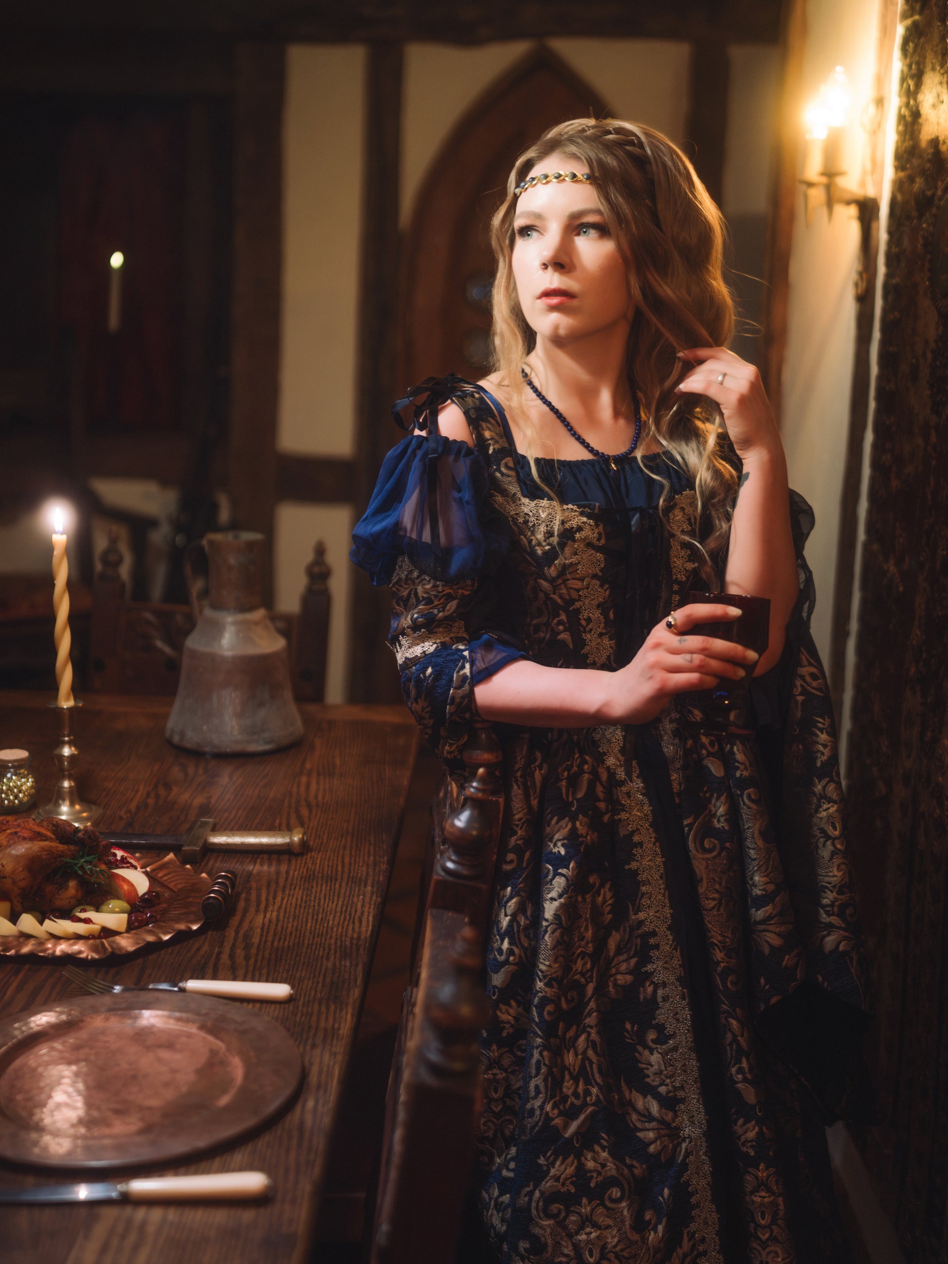 Woman in a medieval-inspired setting wearing a Navy Blue Chenille Corset Dress with gold accents and holding a cup