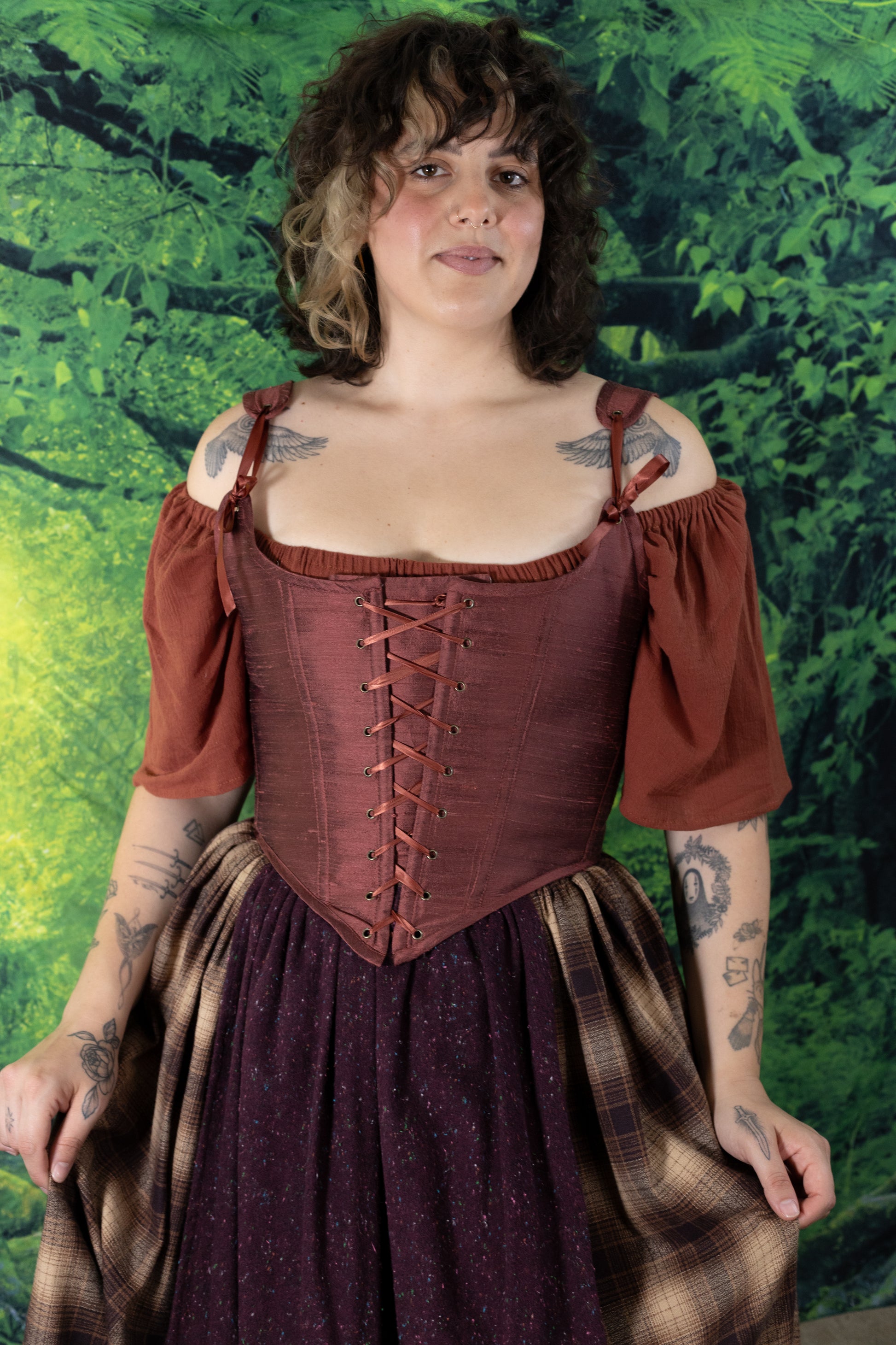 Woman wearing a rust-colored Handmade Mary Sanderson Silk Corset with off-shoulder sleeves and patterned skirt against a green background