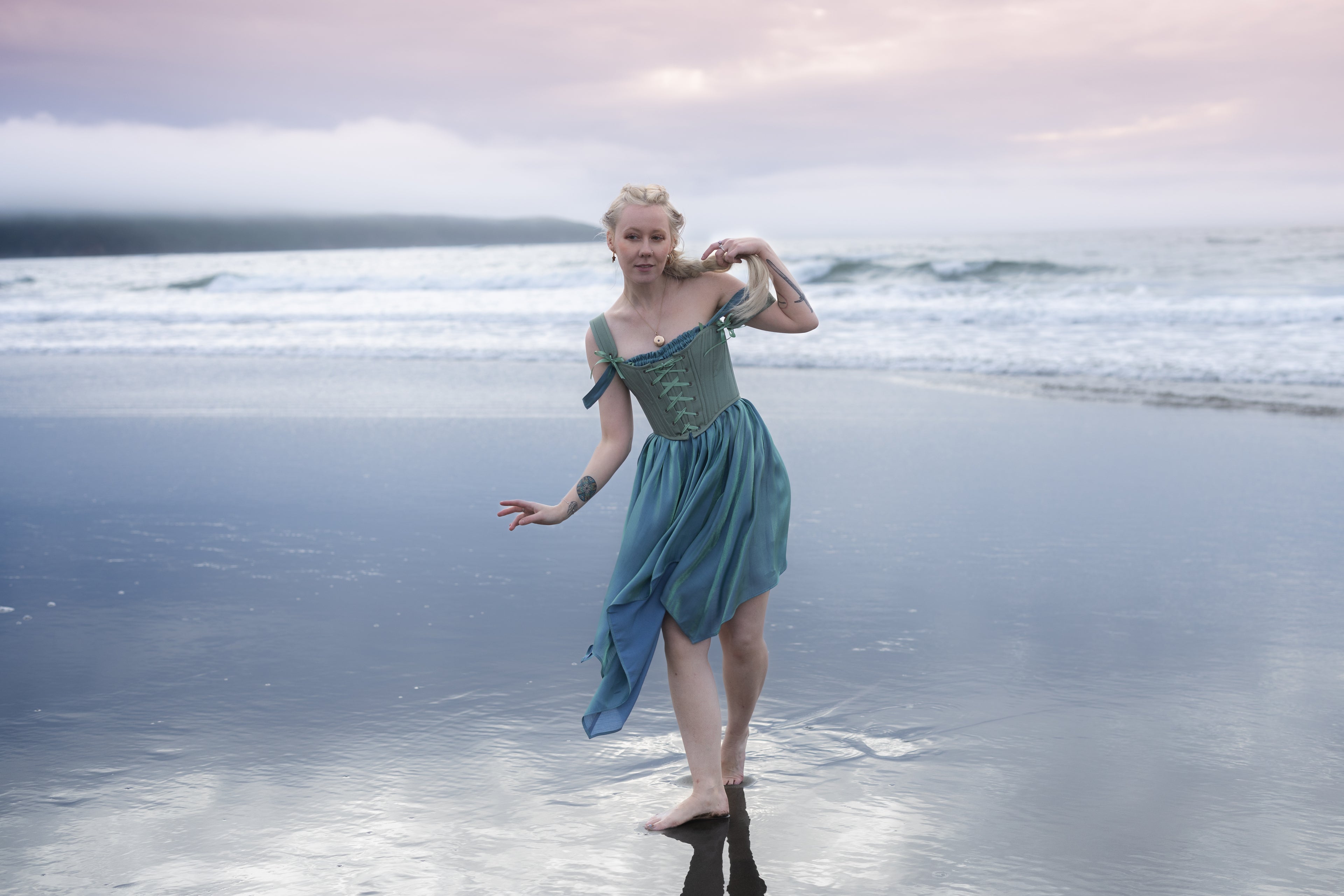 Woman wearing a muted green corset bodice and flowing teal skirt standing barefoot on a wet beach at sunset