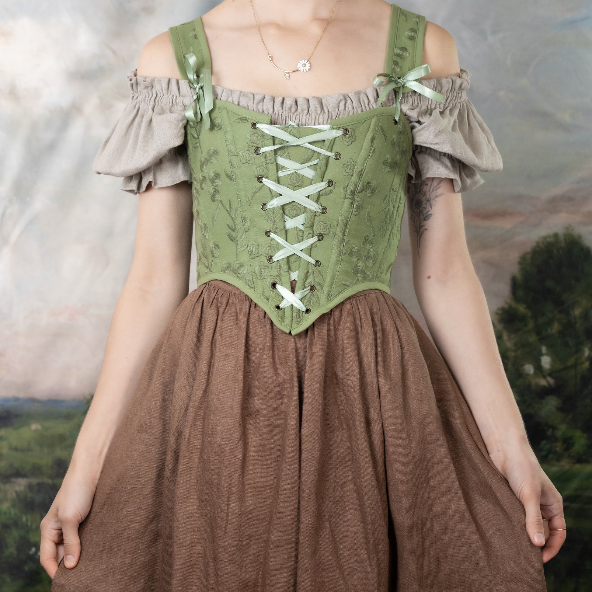 Moss Green Embroidered Floral Corset Top with off-shoulder sleeves, front lacing detail, and brown skirt against pastoral background