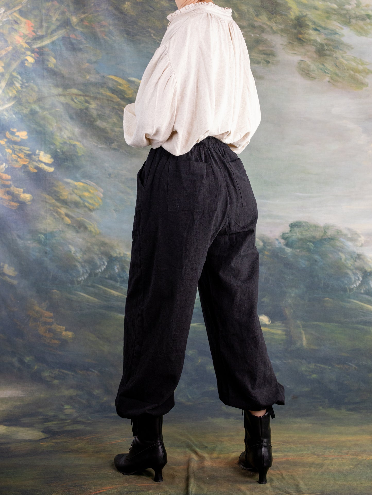 Side view of a model wearing the Borgia Shirt with loose black pants and black boots against a scenic backdrop