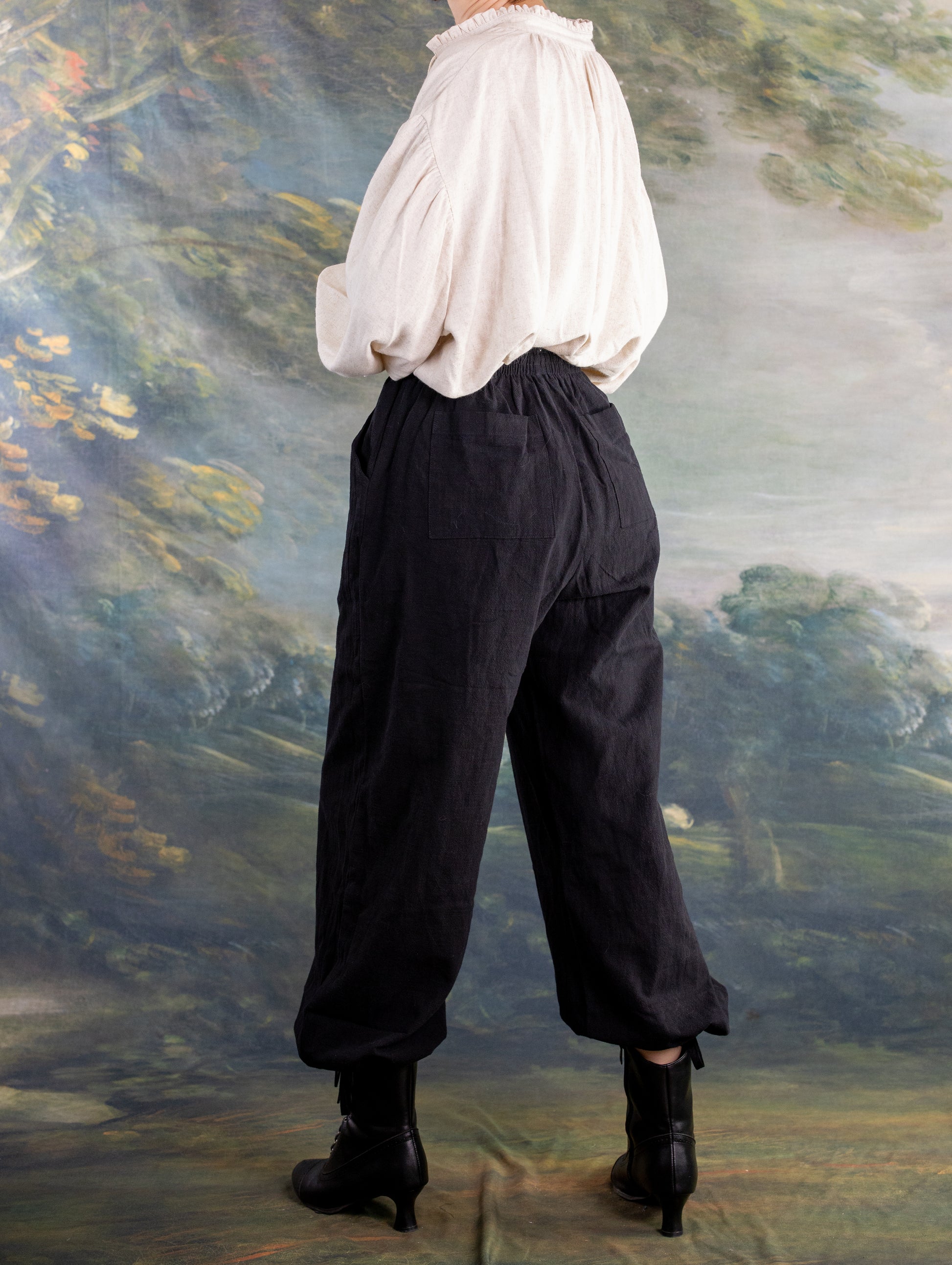 Side view of a model wearing the Borgia Shirt with loose black pants and black boots against a scenic backdrop