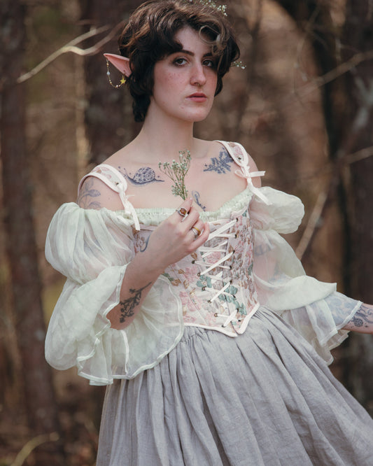 Young person with elf ears in a vintage corset dress standing in a forest holding a small flower