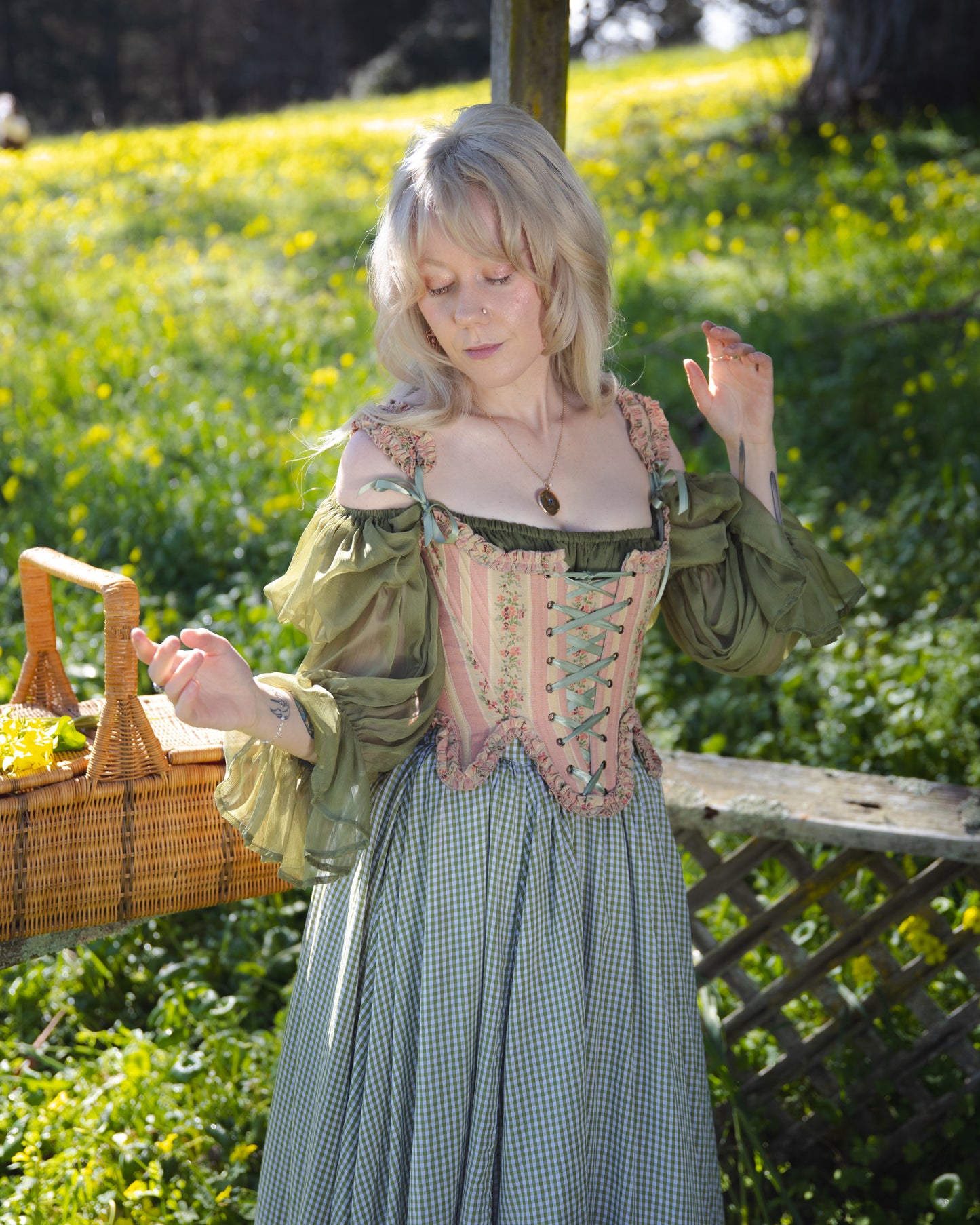 Woman wearing Rococo Pink 18th Century Stays corset over a green blouse in a sunlit garden setting.