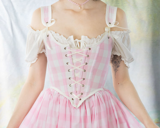 Woman wearing a Handmade Pink Gingham Corset with ruffled off-shoulder blouse and necklace, soft pastel background.
