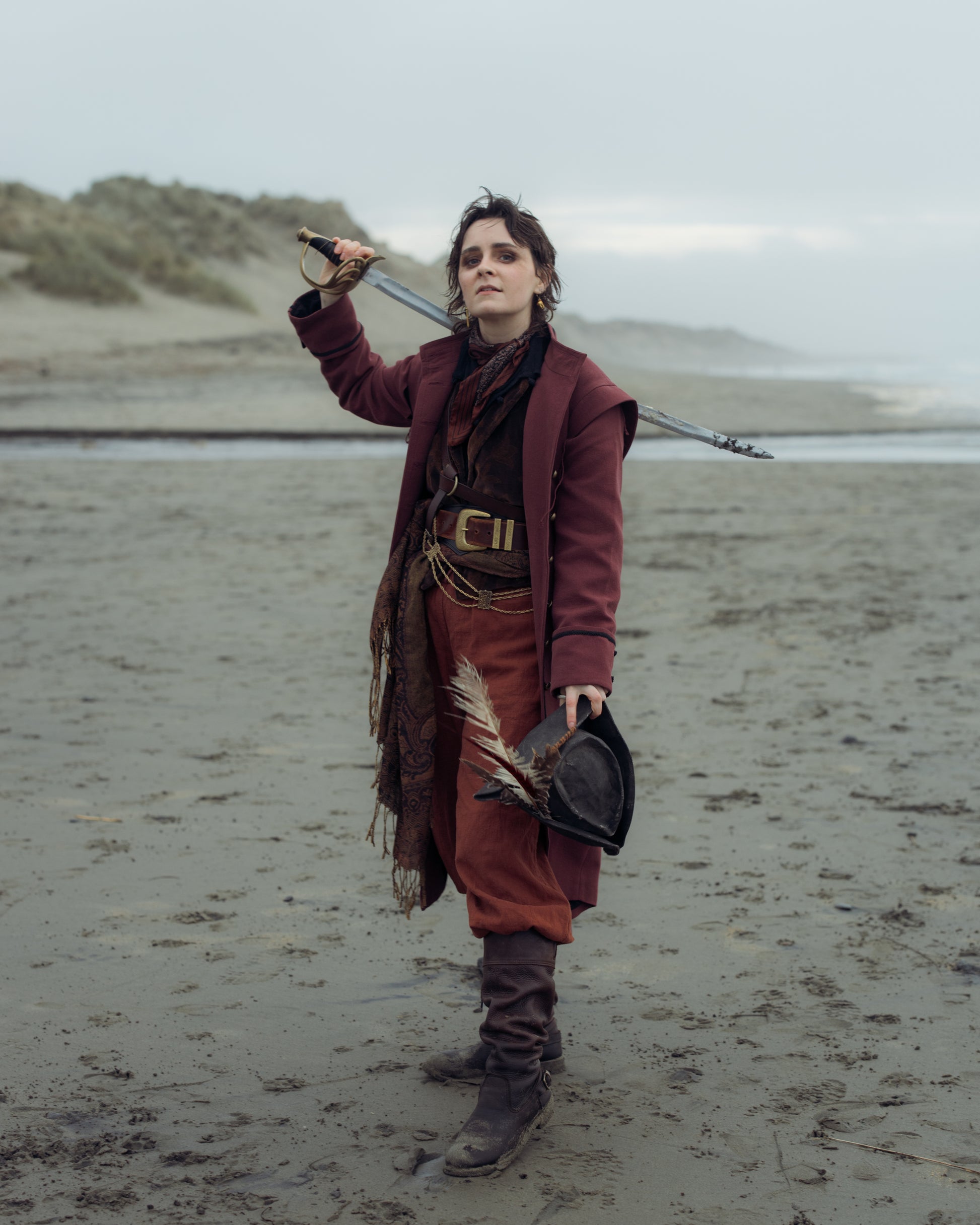 Person standing on a beach wearing the Pirate Captain's Coat holding a sword over the shoulder and a feathered hat in hand