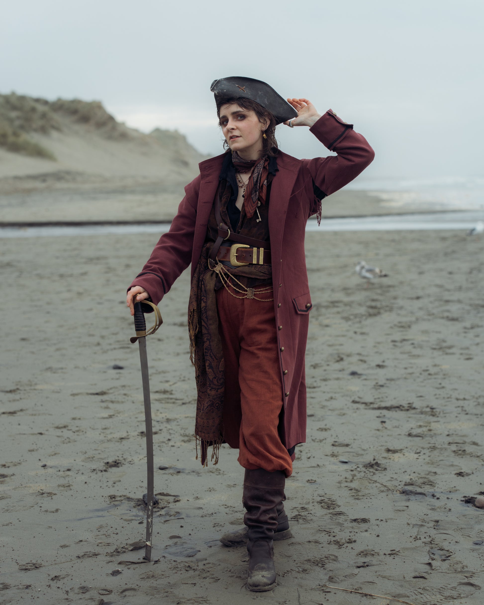 Person wearing a Pirate Captain's Coat and hat standing on a beach holding a cutlass sword with sand dunes in background