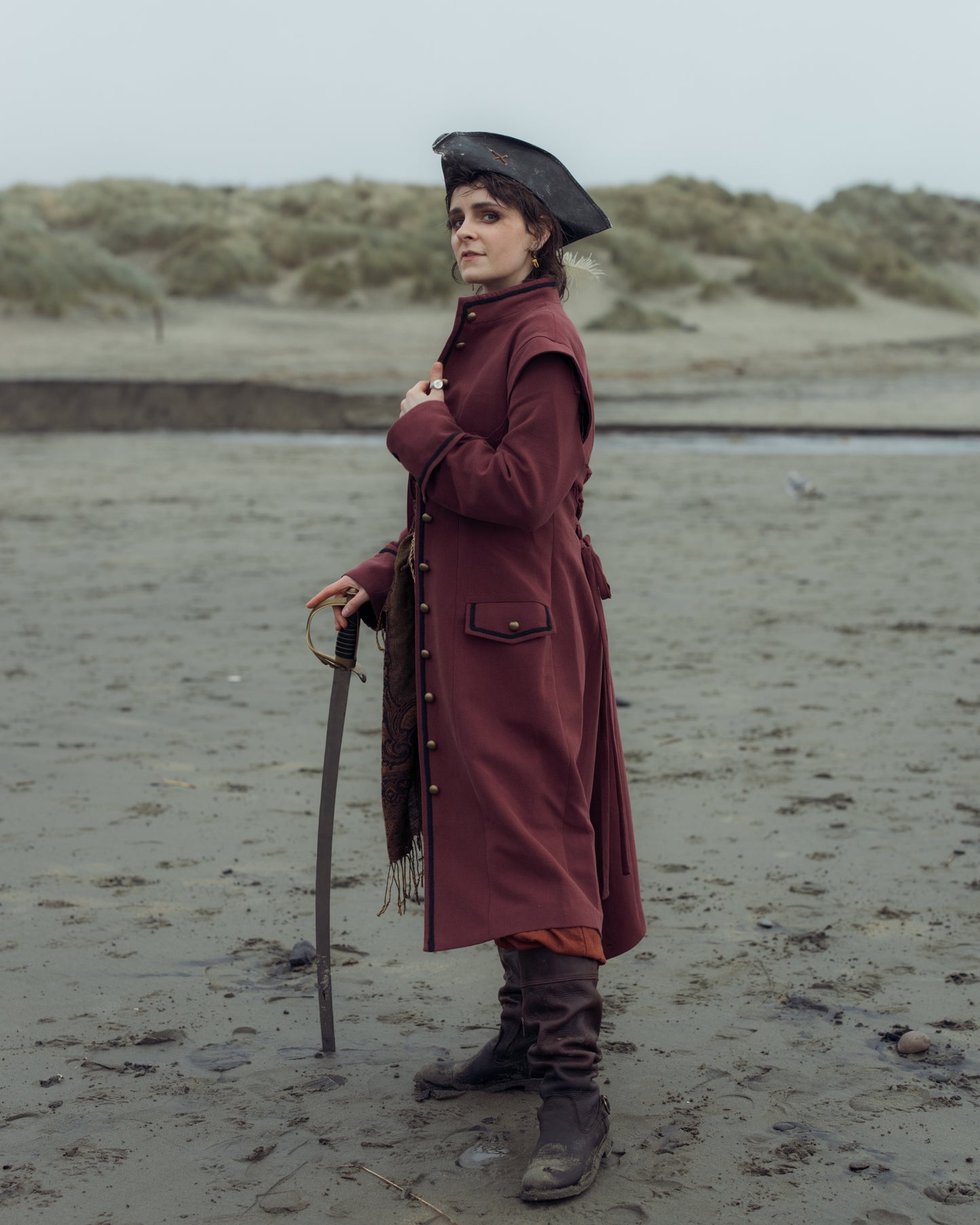 Person standing on a beach wearing the Pirate Captain's Coat with a tricorn hat and holding a sword