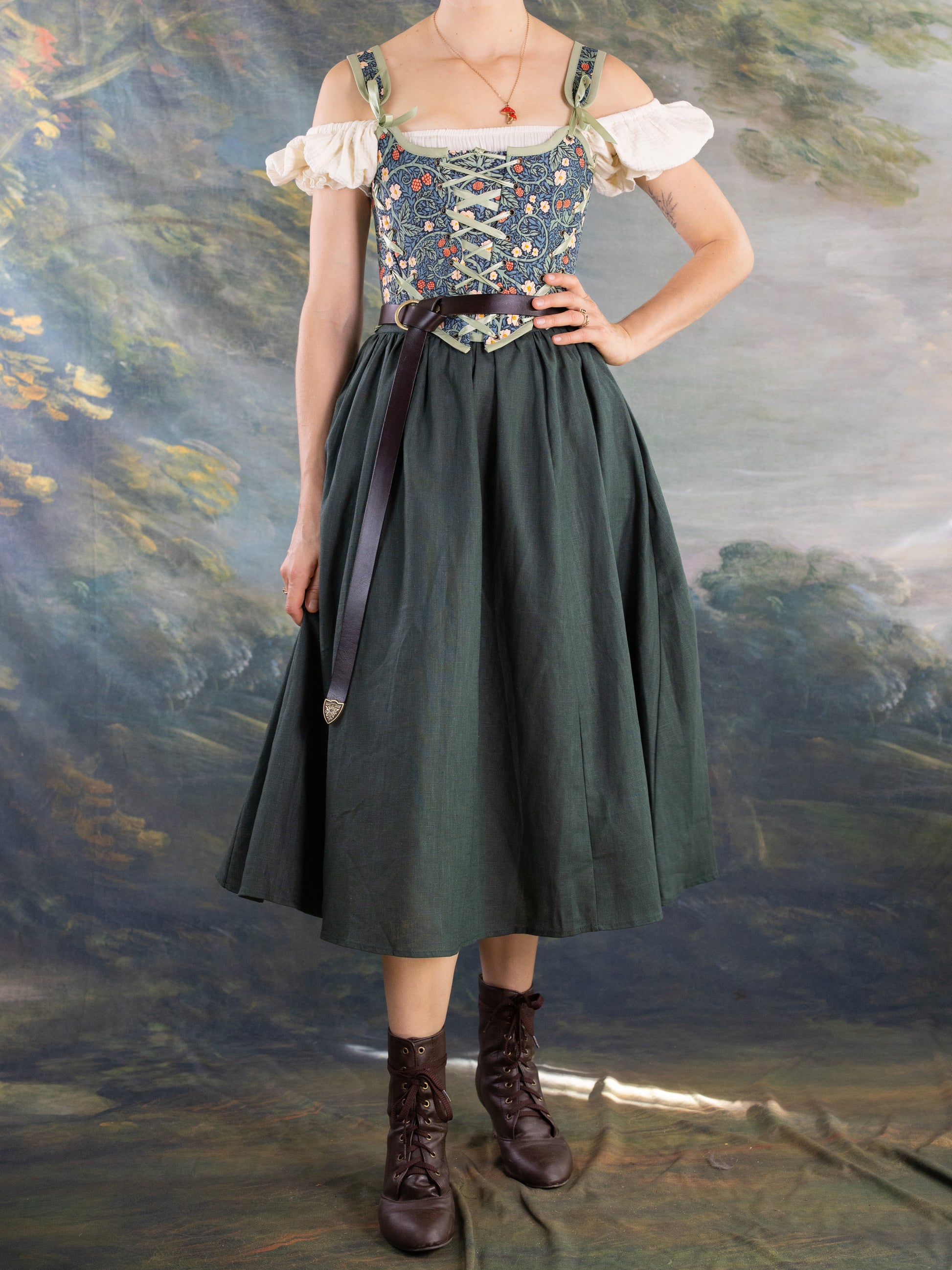 Woman wearing a floral Raspberry Vines Princess Stays corset with off-shoulder blouse and dark green skirt standing in front of painted backdrop
