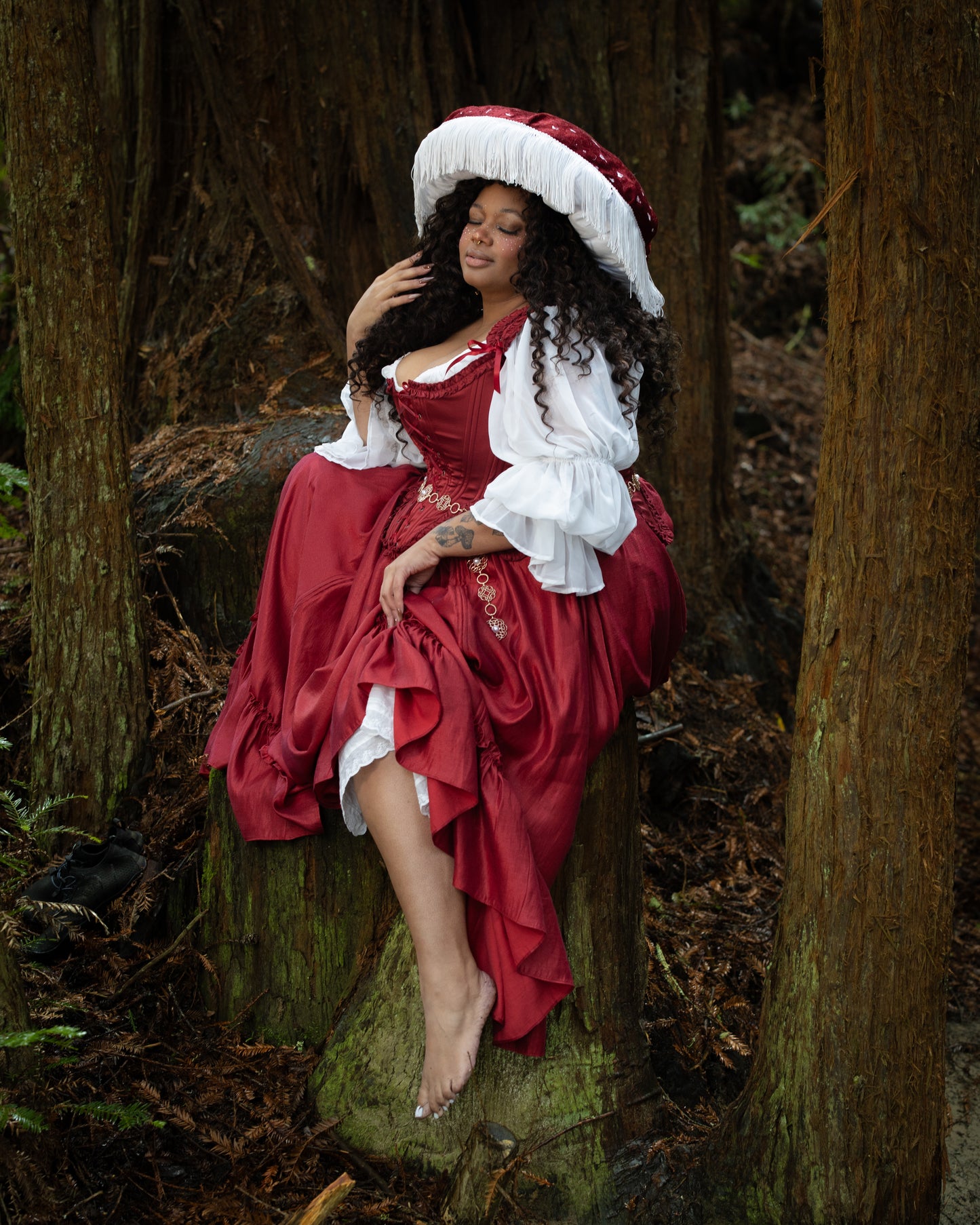 Model seated on tree stump in gallery position 5 wearing dark red ruffle skirt with elegant rococo styling outdoors