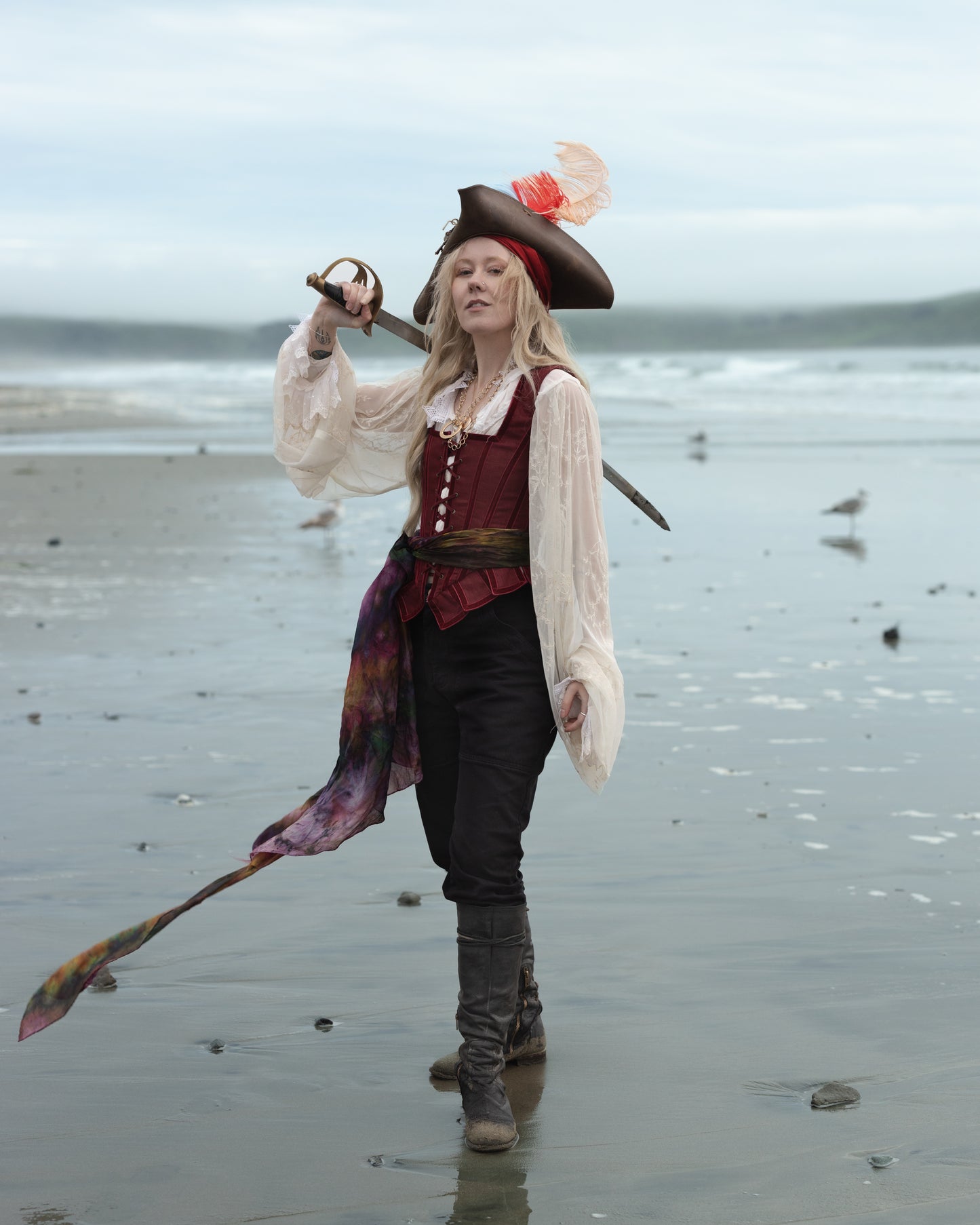 Woman standing on beach wearing Red Pirate Doublet with sword and feathered hat in foggy seaside setting