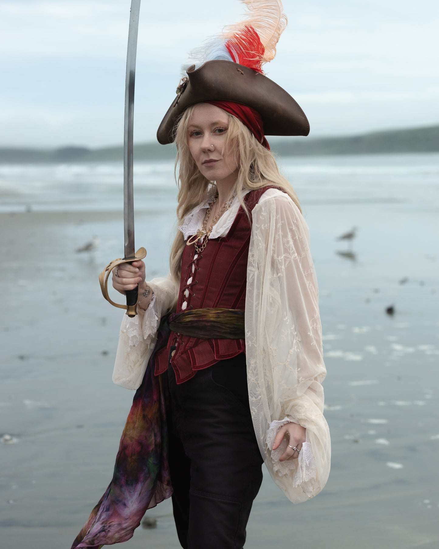 Woman wearing Red Pirate Doublet costume holding a sword with ocean and seagulls in the background