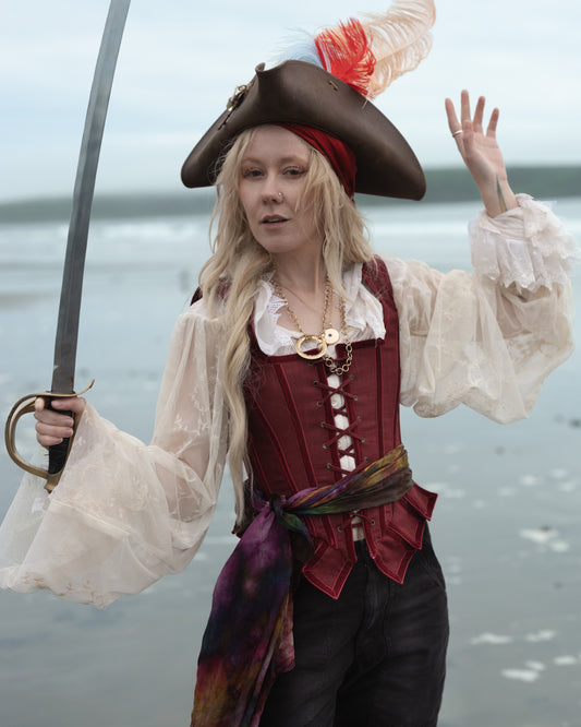 Woman on beach wearing Red Pirate Doublet with billowy white sleeves, feathered hat, and holding a sword raised in the air