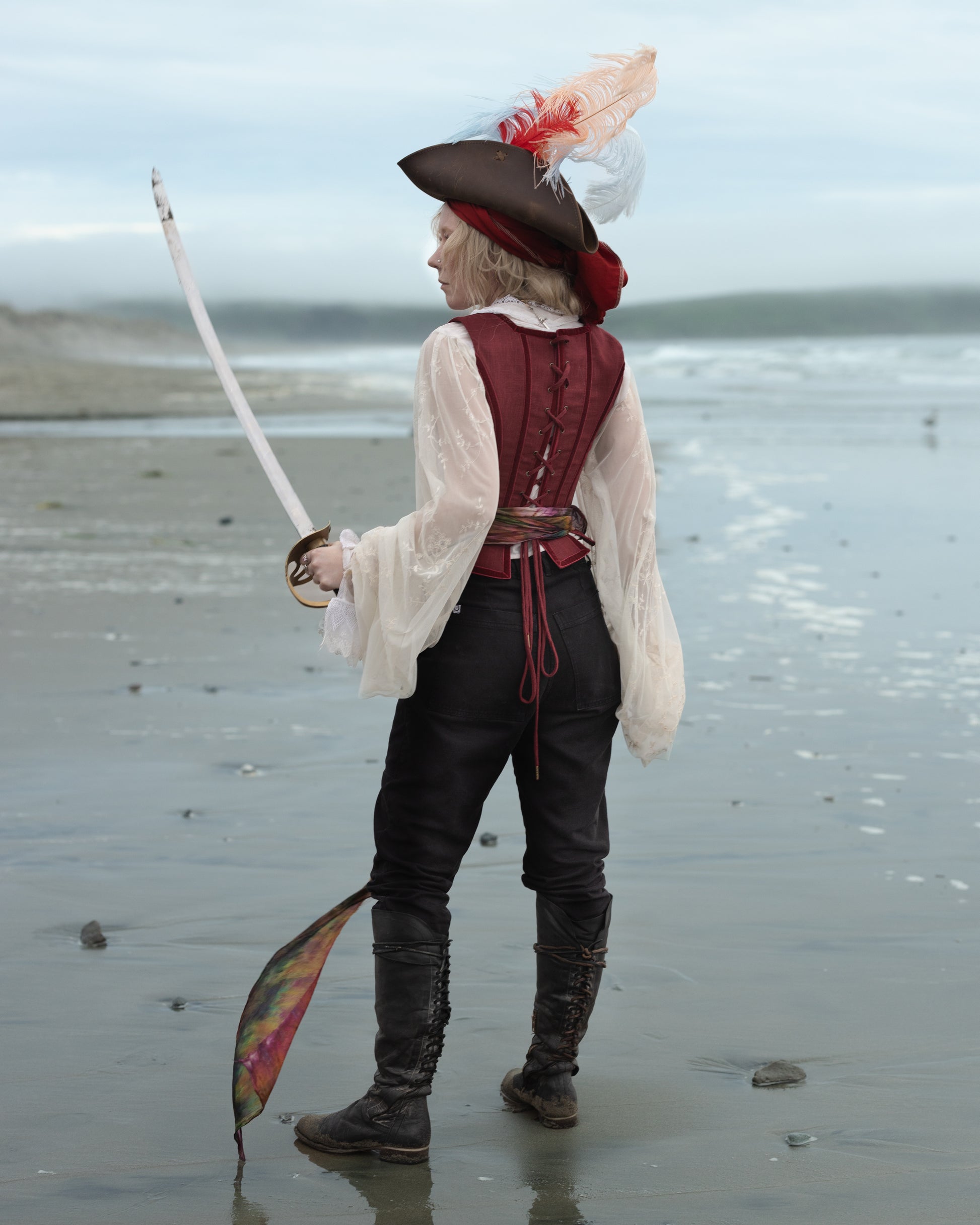Person in Red Pirate Doublet holding a sword on a beach with a feathered pirate hat and tall boots