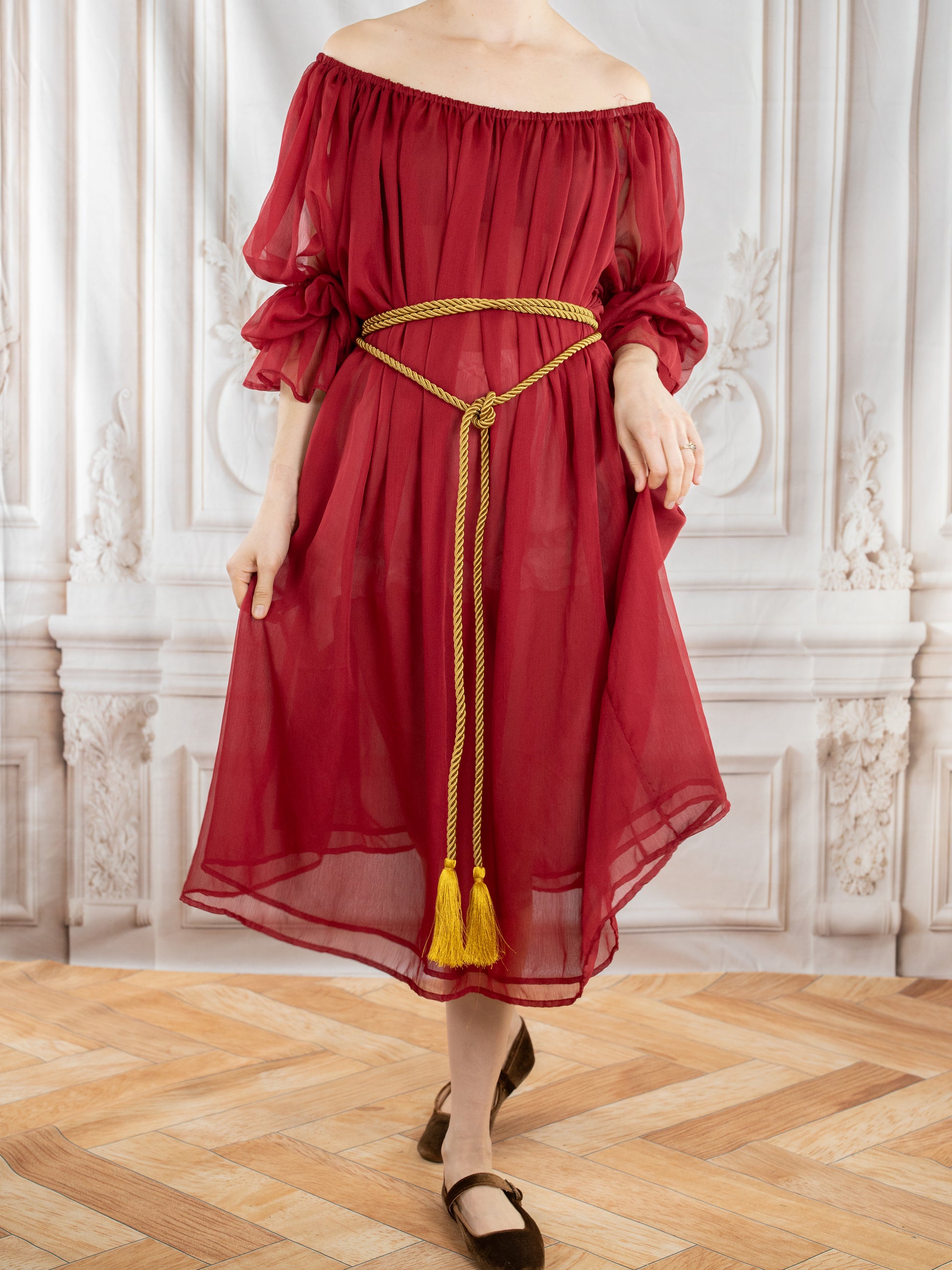 Woman wearing a flowing Red Chiffon Chemise with off-shoulder design and gold tasseled belt in elegant room.