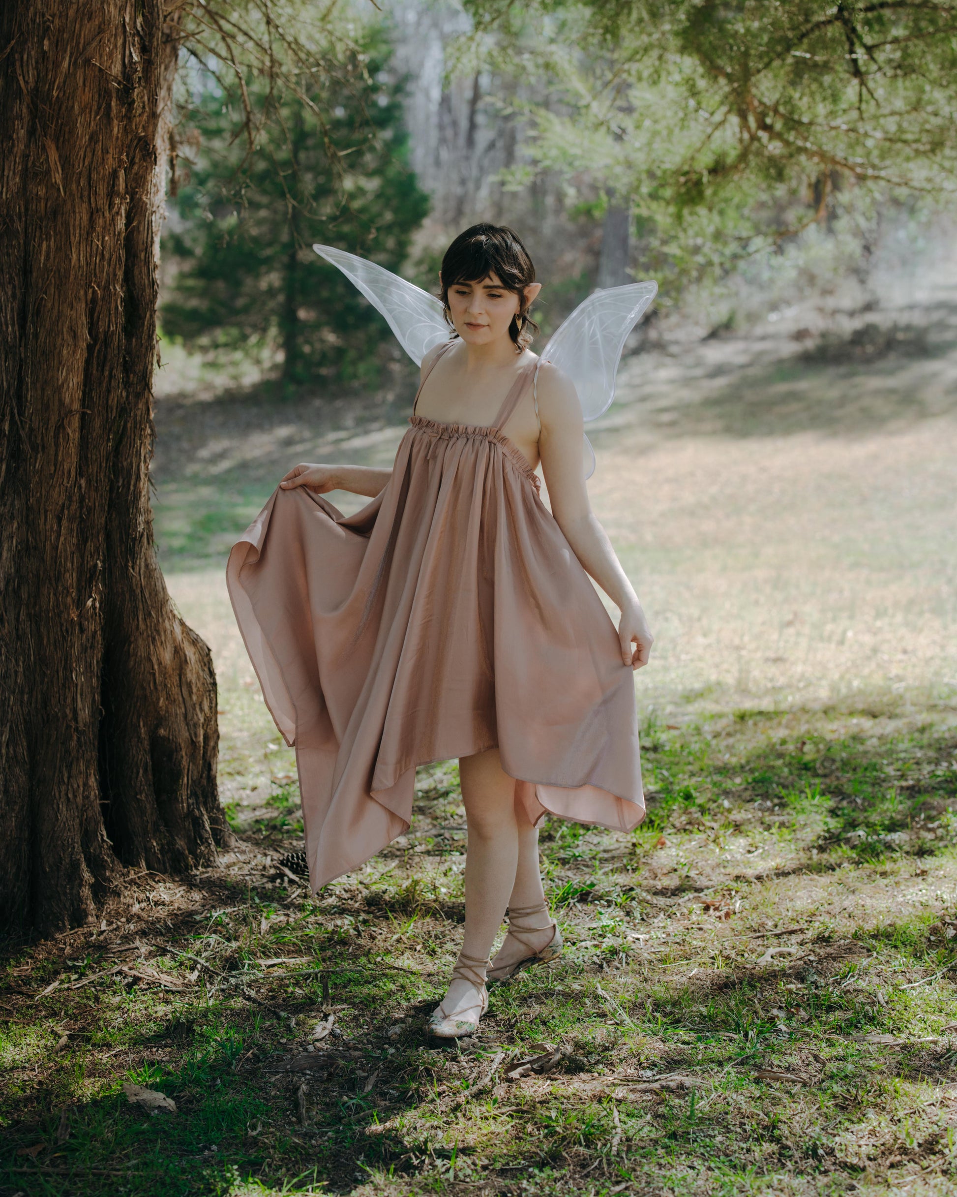 Young woman with light wings wearing a flowing dress standing near a tree in a sunlit forest clearing