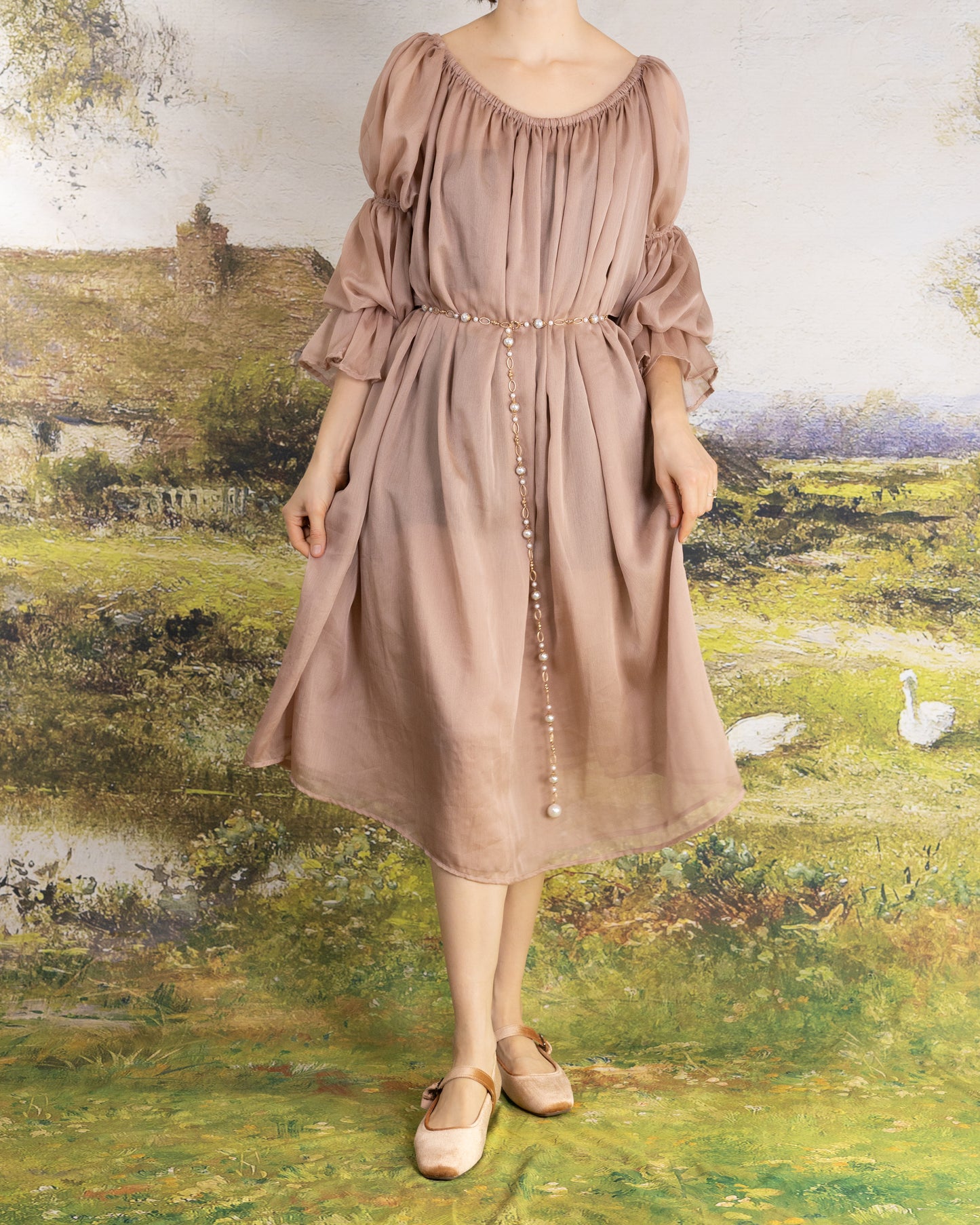 Woman wearing Rose Chiffon Chemise in muted rose with puff sleeves and pearl belt, standing in a scenic garden setting