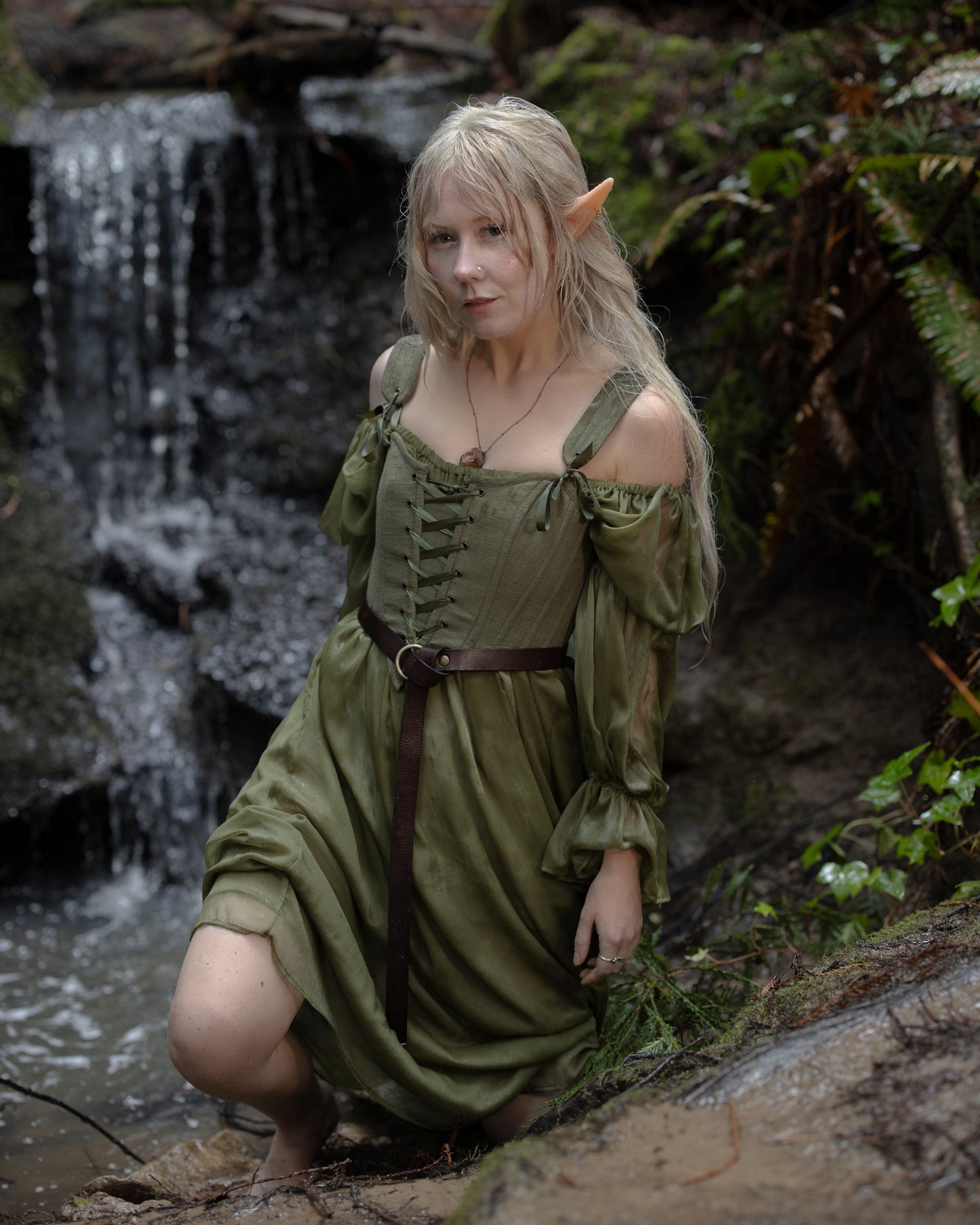 Woman wearing Sage Cotton Corset with off-shoulder sleeves and belt, seated near a waterfall in a forest setting