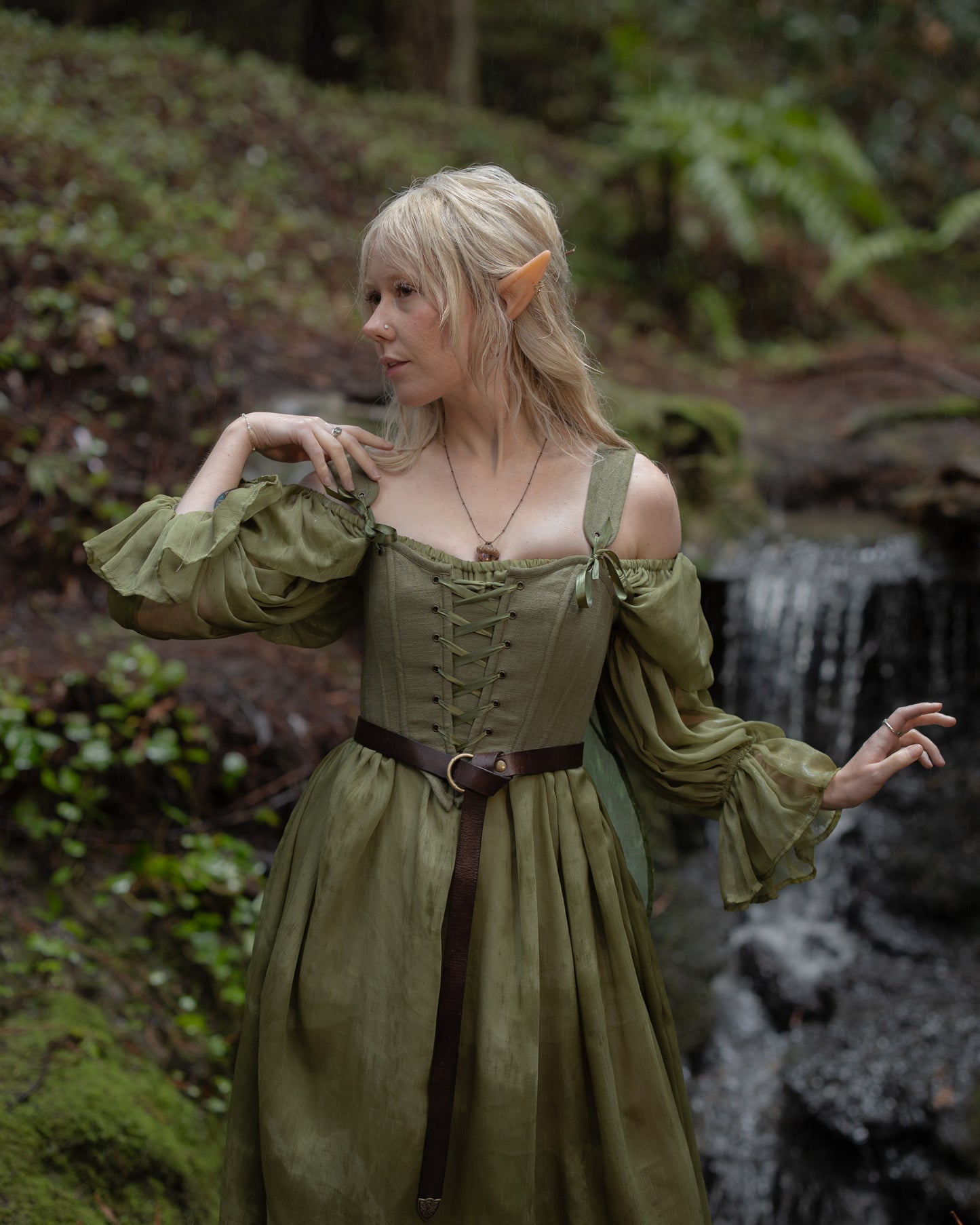 Woman in a forest wearing a lace-up Sage Cotton Corset with off-shoulder sleeves and a brown belt standing near a waterfall