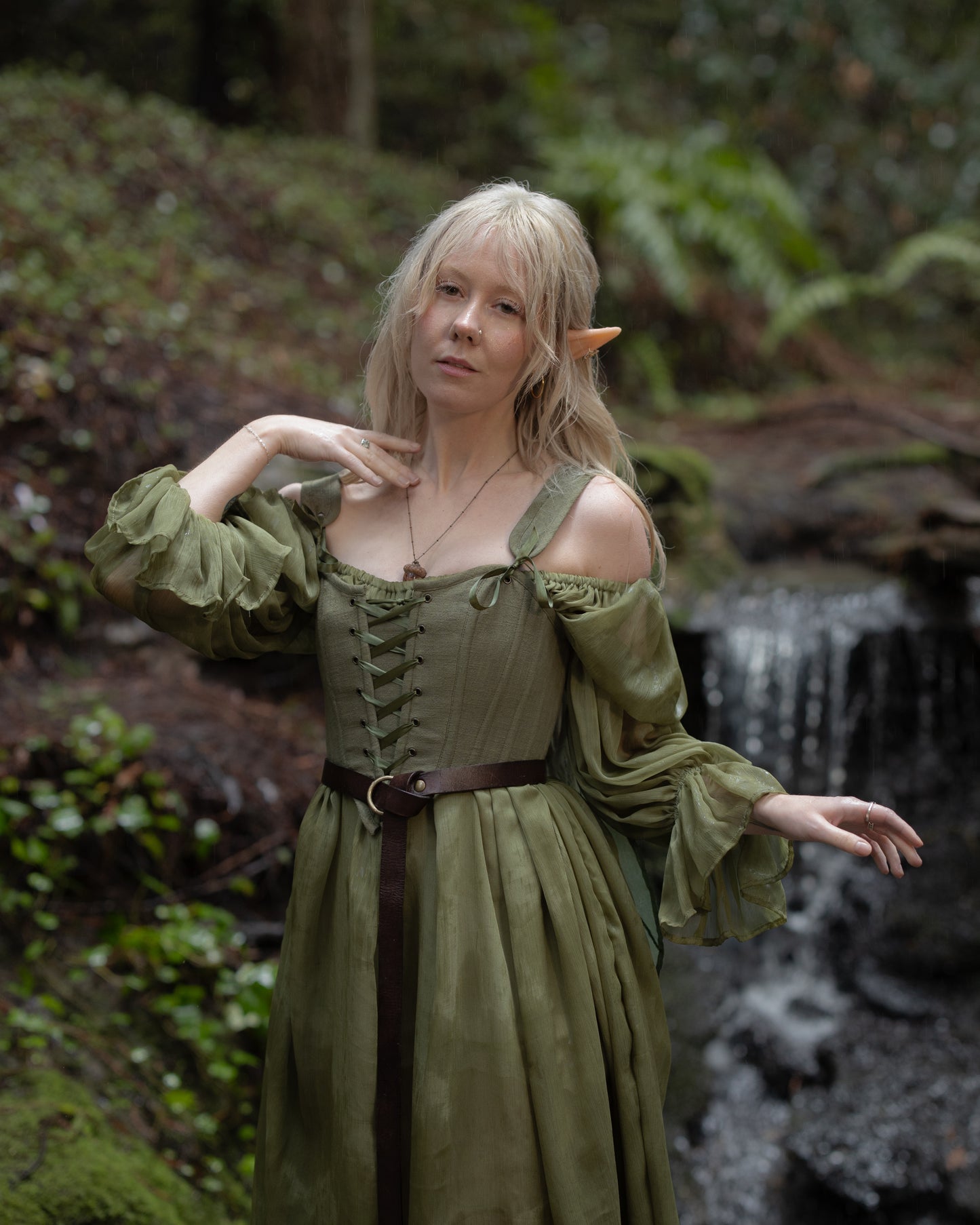 Woman wearing a Sage Cotton Corset with lace-up front and green flowing sleeves standing near a forest stream