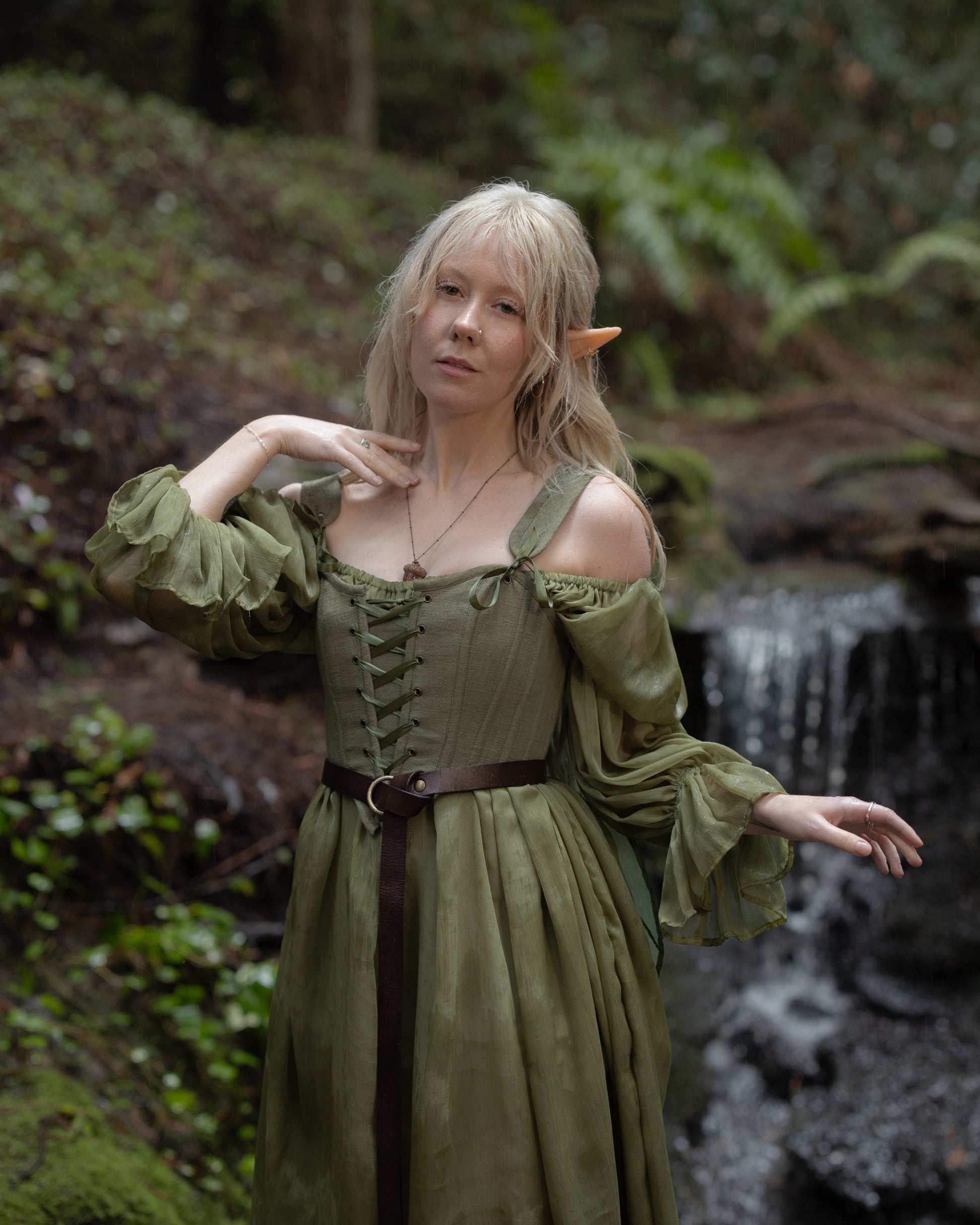 Woman wearing a Sage Cotton Corset with lace-up front and green flowing sleeves standing near a forest stream
