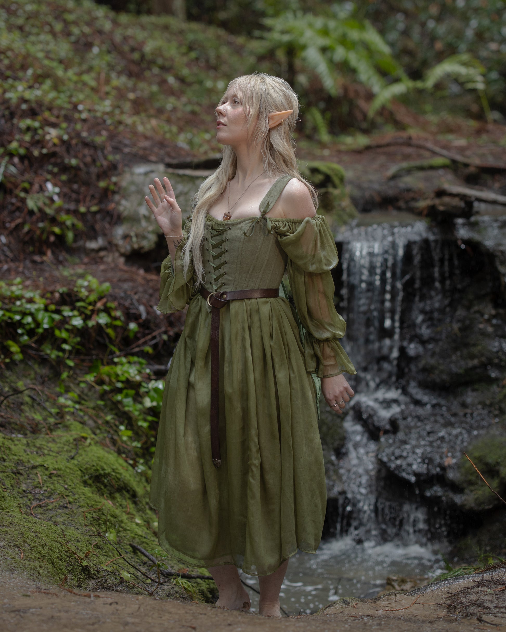 Woman in forest wearing earthy green Sage Cotton Corset with flowing skirt and sheer sleeves beside waterfall