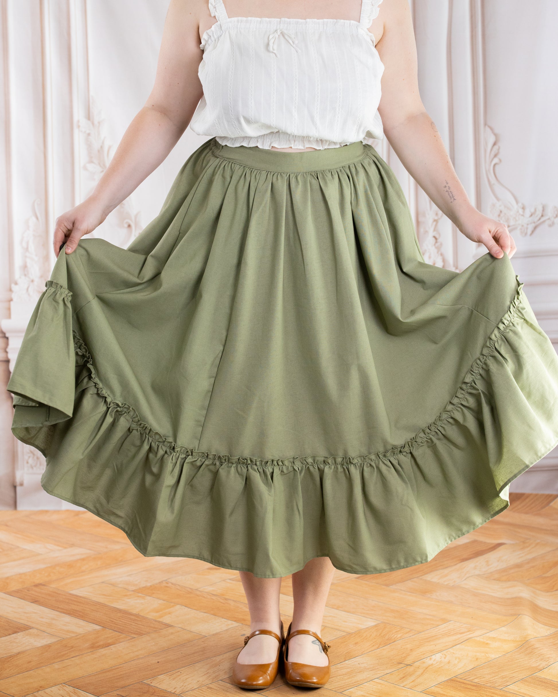 Woman wearing Sage Linen Ruffle Skirt in sage green holding out skirt sides indoors on wooden floor background