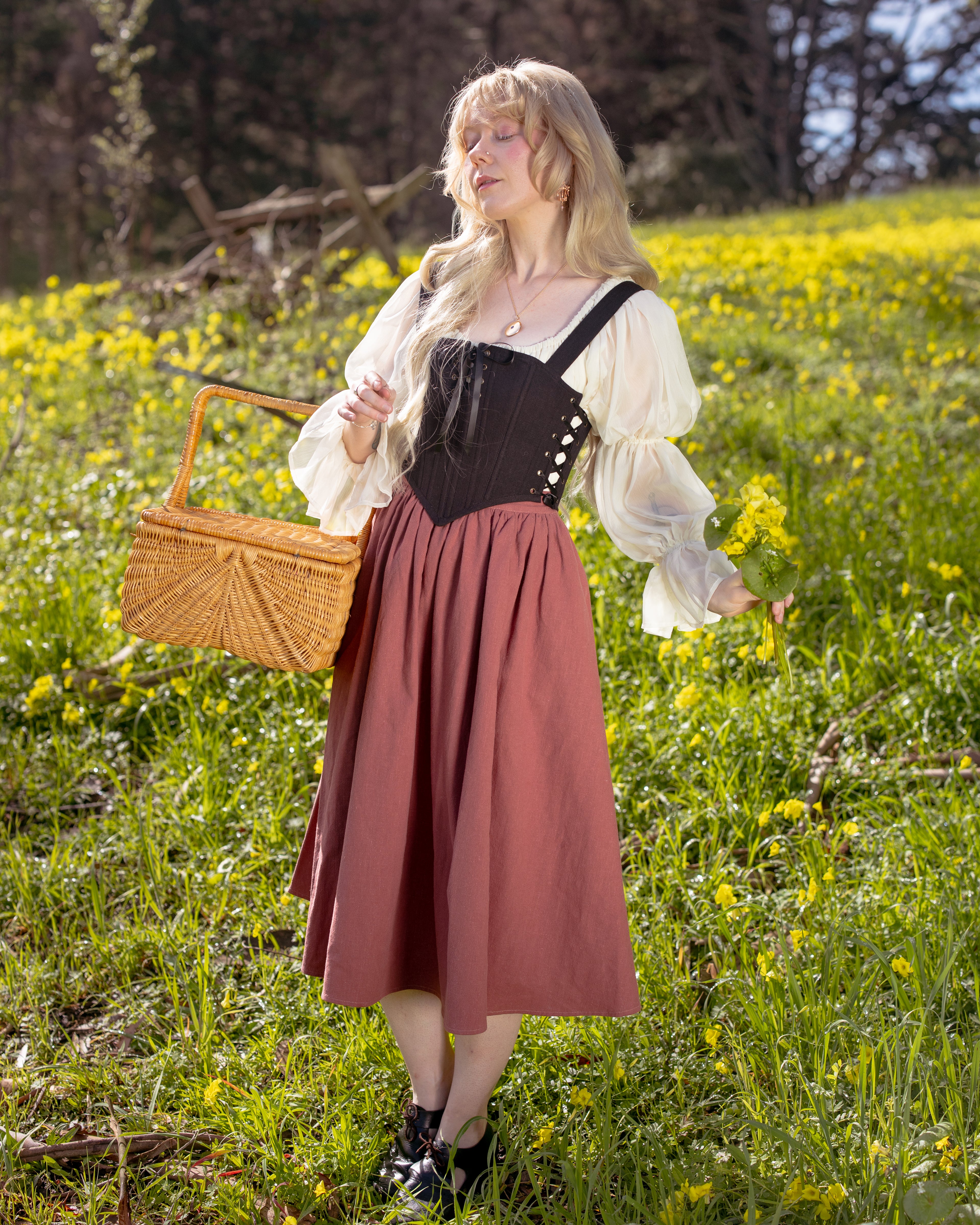 Woman wearing Black Aurora Stays corset over cream blouse and rust skirt standing in sunny flower field holding a basket and flowers