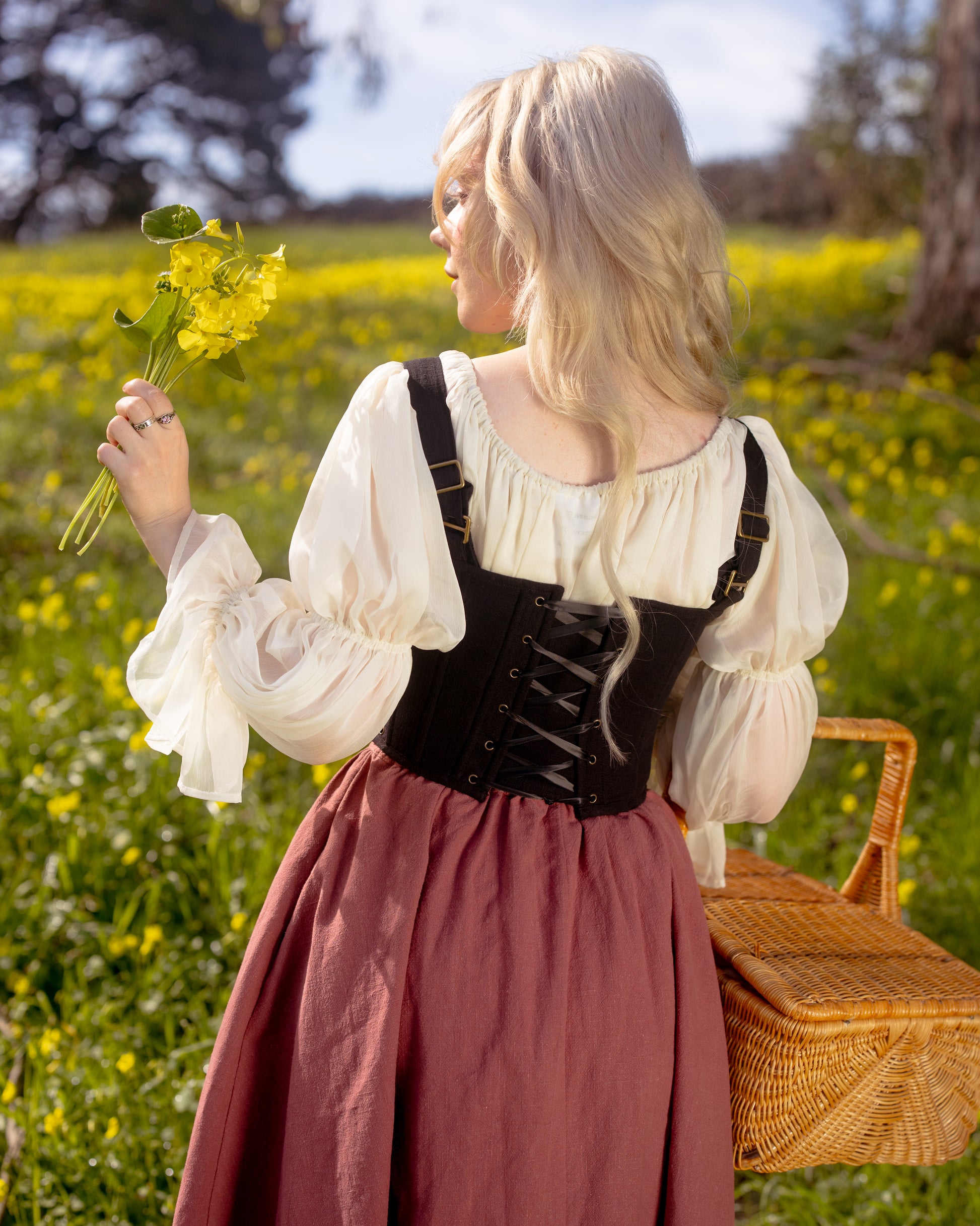 Woman in countryside wearing Black Aurora Stays corset over white blouse holding yellow flowers by wicker basket
