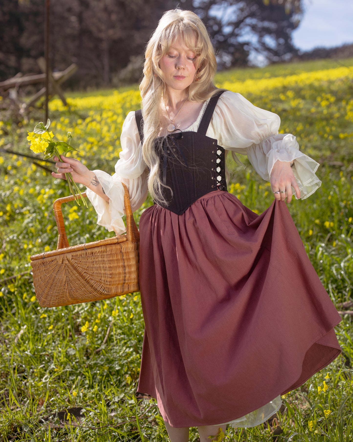 Woman in flowing dress holding a basket and flowers in a sunny field wearing Black Aurora Stays corset top