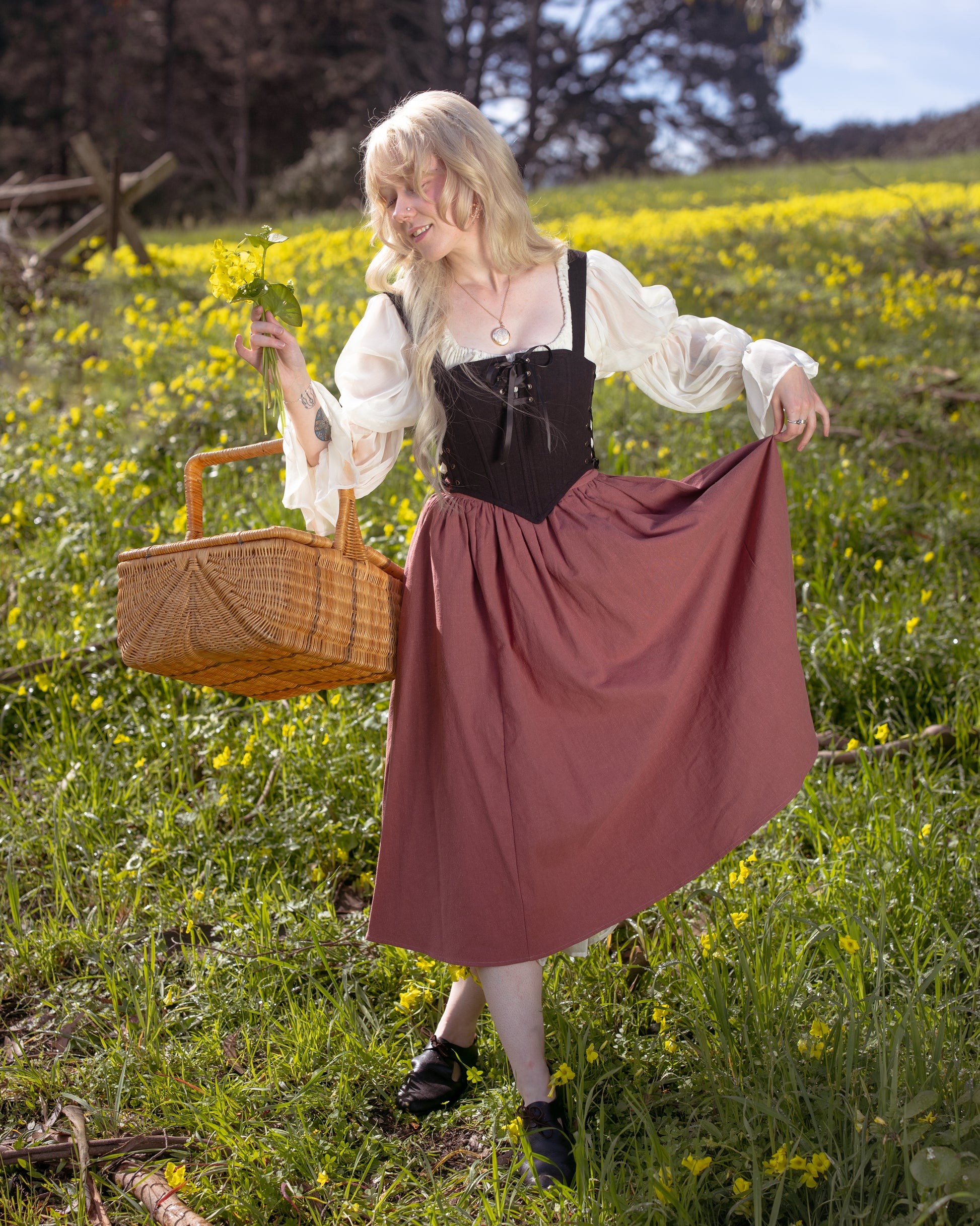 Woman in a flowing dress and Black Aurora Stays corset holding flowers and a wicker basket in a sunny field of yellow blooms