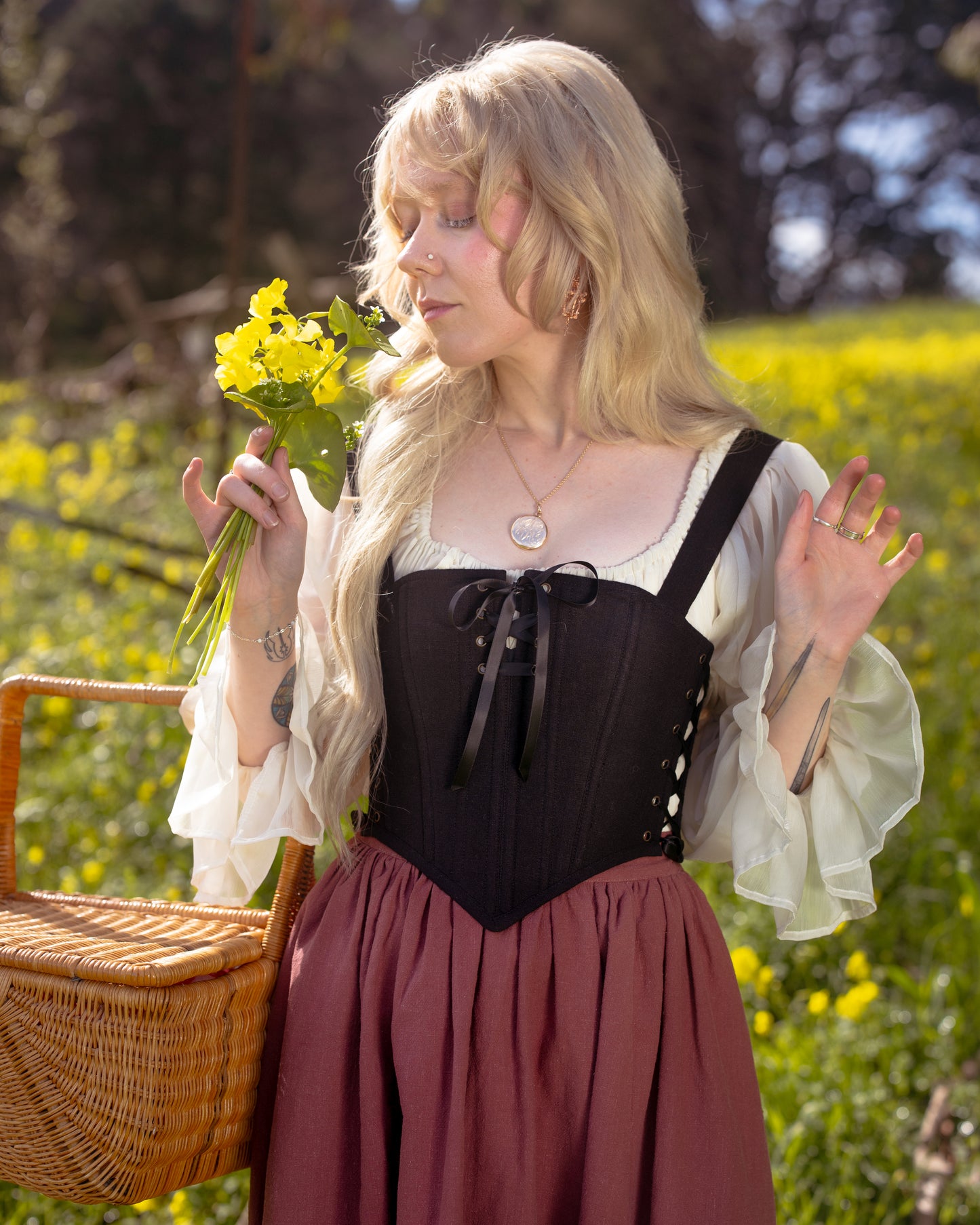 Woman wearing Black Aurora Stays corset over white blouse holding yellow flowers in a sunny field with a wicker basket nearby