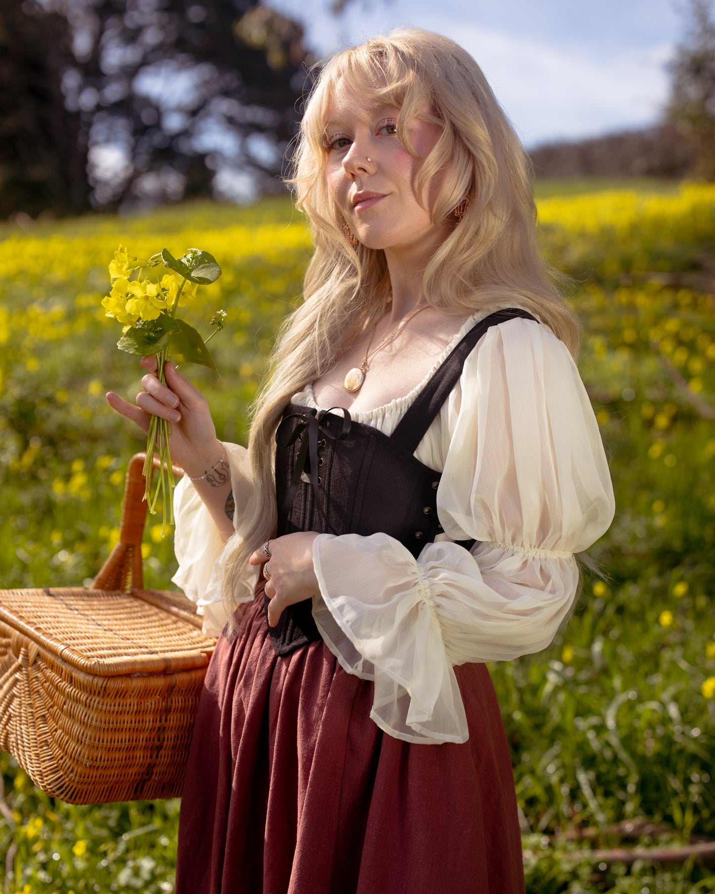 Woman in a flowing blouse and skirt holding flowers and a basket wearing Black Aurora Stays corset outdoors in a field
