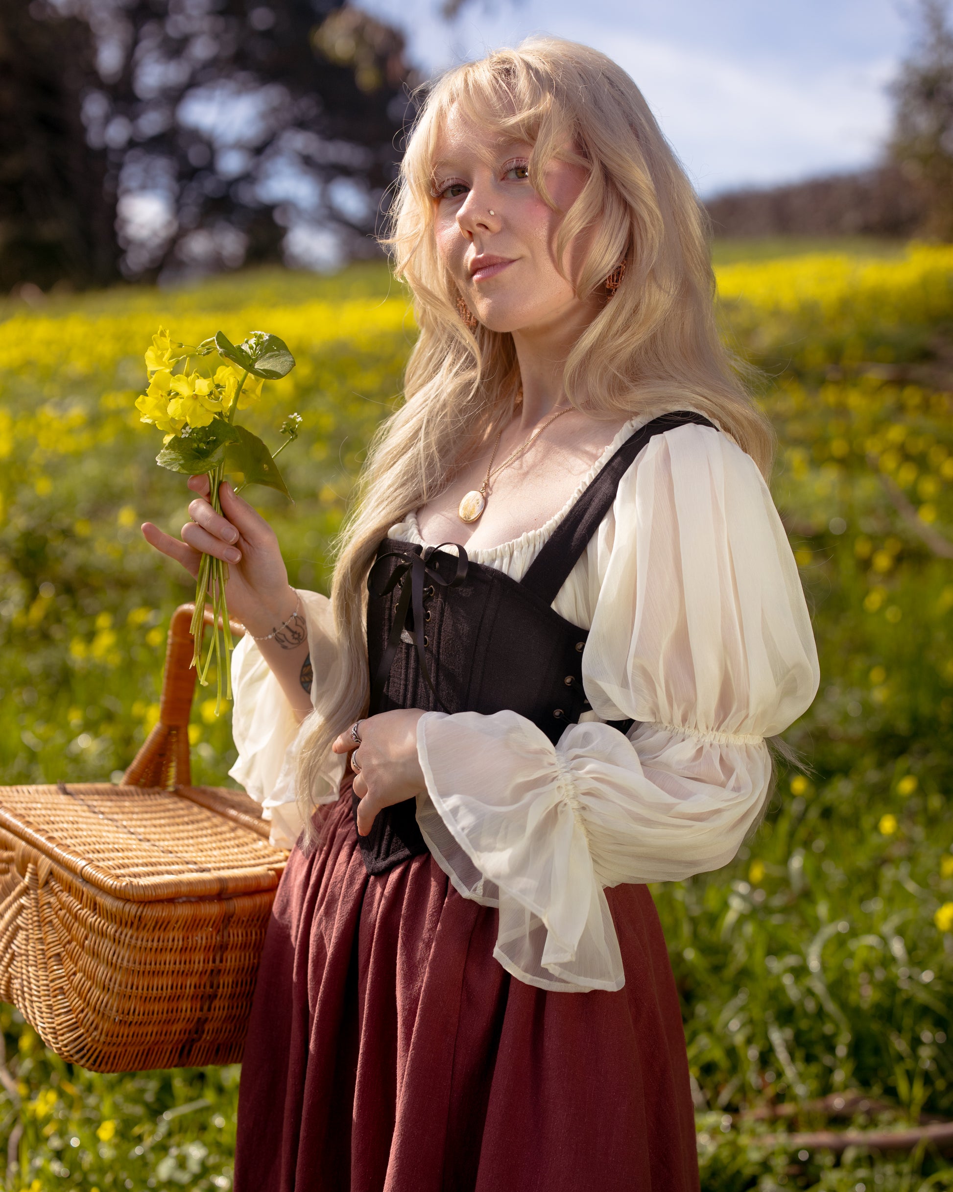 Woman in a flowing blouse and skirt holding flowers and a basket wearing Black Aurora Stays corset outdoors in a field