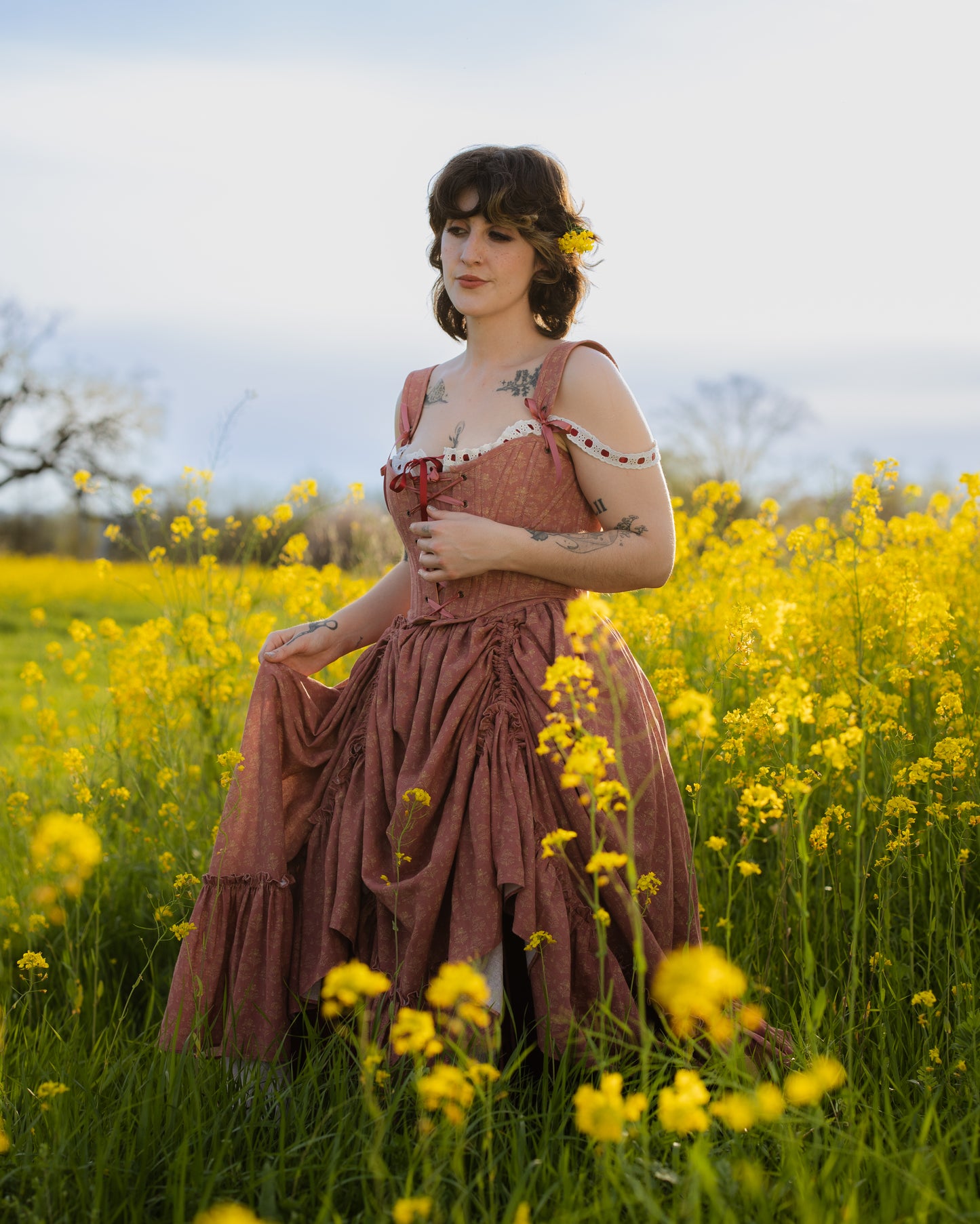 Woman wearing a Strawberry Floral Regency Stays dress standing in a field of yellow wildflowers with a flower in her hair