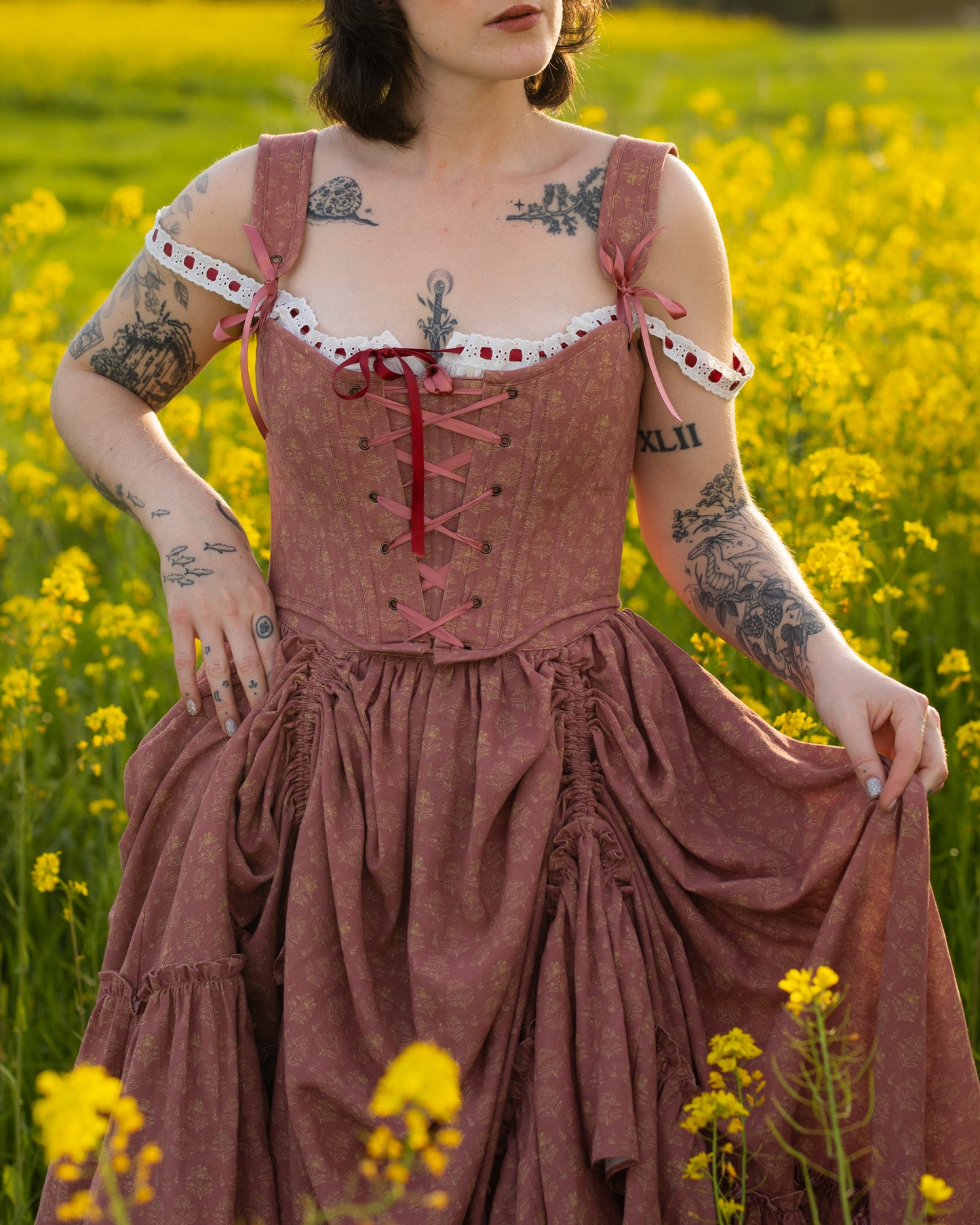 Woman wearing a pink dress with lace and ribbon details in a yellow flower field, showcasing Strawberry Floral Regency Stays