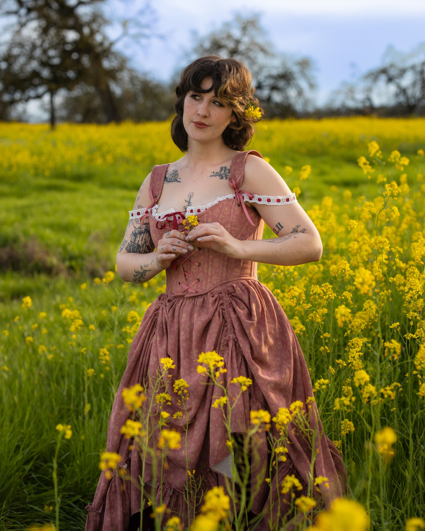Woman wearing Strawberry Floral Regency Stays dress stands in a yellow flower field holding small blossoms.
